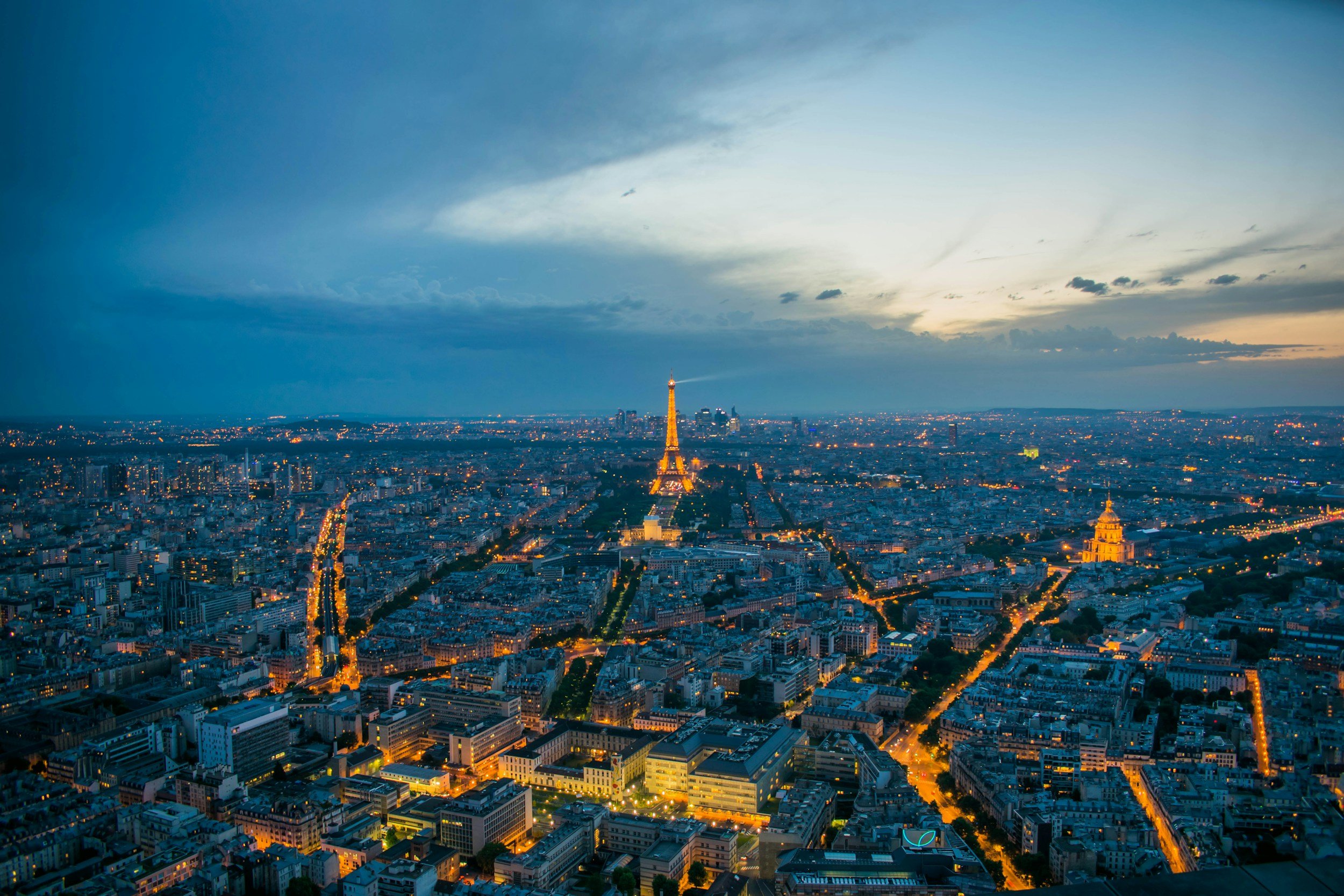 Aerial view of Paris at dusk, featuring the illuminated Eiffel Tower and city streets with glowing lights.
