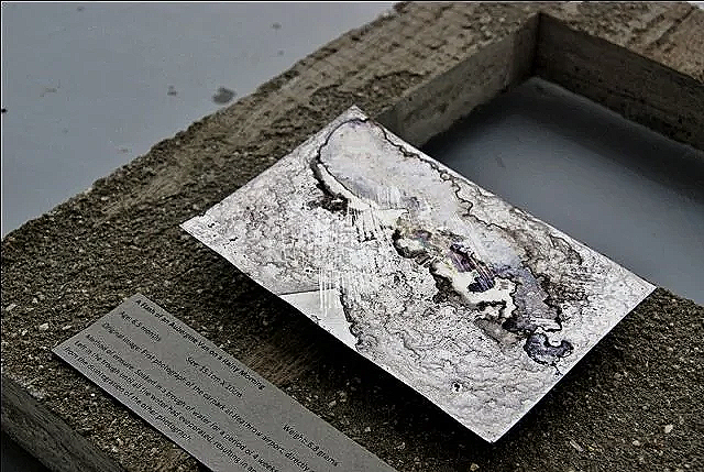 A disintegrated photo print with its image barely discernible, much of it is covered in grey dust, leaving only the sky a pale lilac blue.