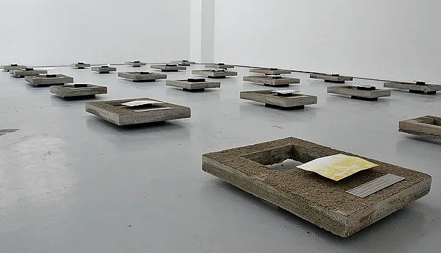 Installation view of the exhibition in which disintegrated photo prints are displayed close to the ground, each lying on a concrete slab.