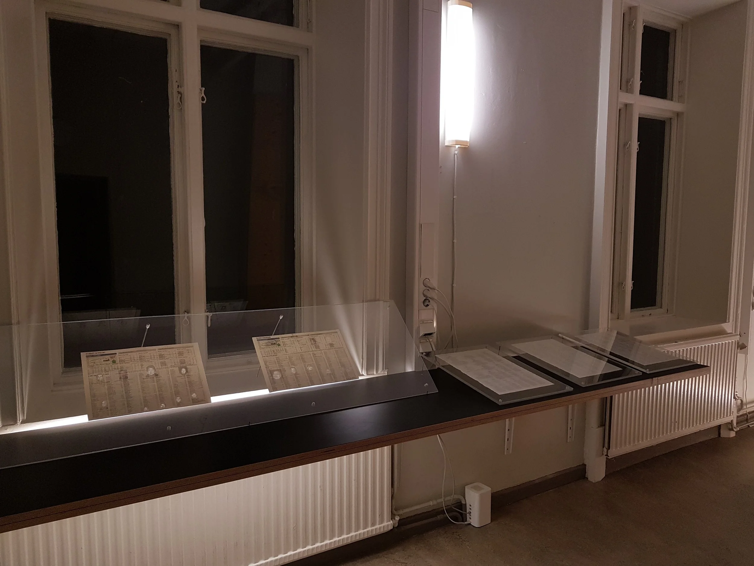 Night-time view of the left-hand side of the room with pages from the Straits Times newspaper displayed in plexi glass frames and lit by a light box.