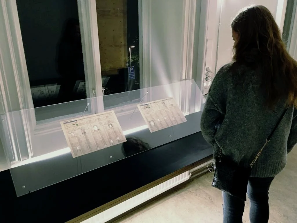 Female visitor with long dark hair in back view looking to the left at the illuminated panel of newspaper prints.