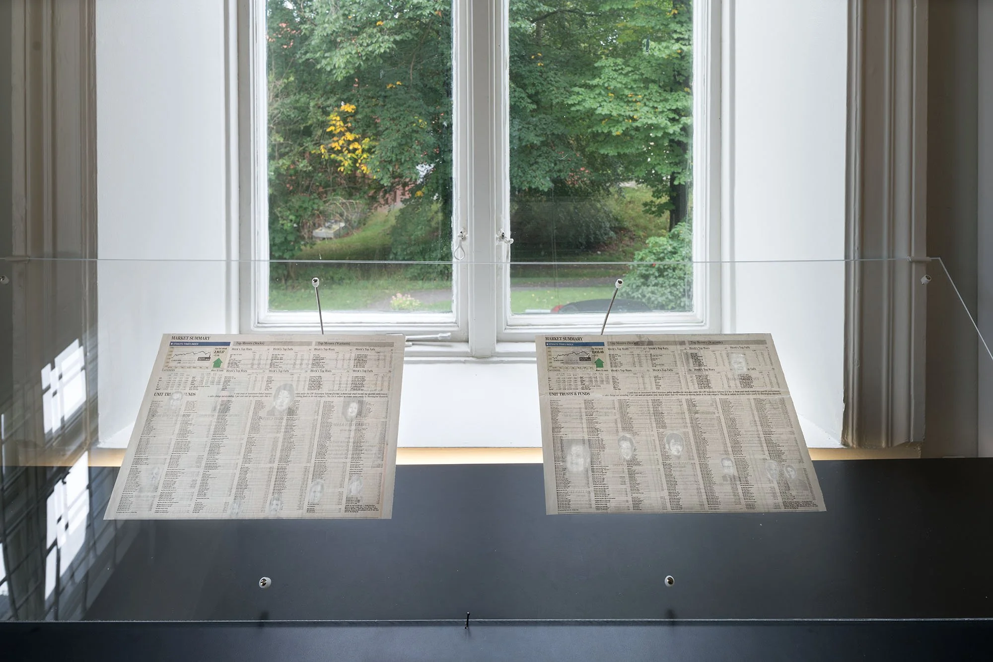 Day-time view of the left-hand side of the gallery with two thin sheets of newspaper framed in a clear plexi glass sheet in front of a window.