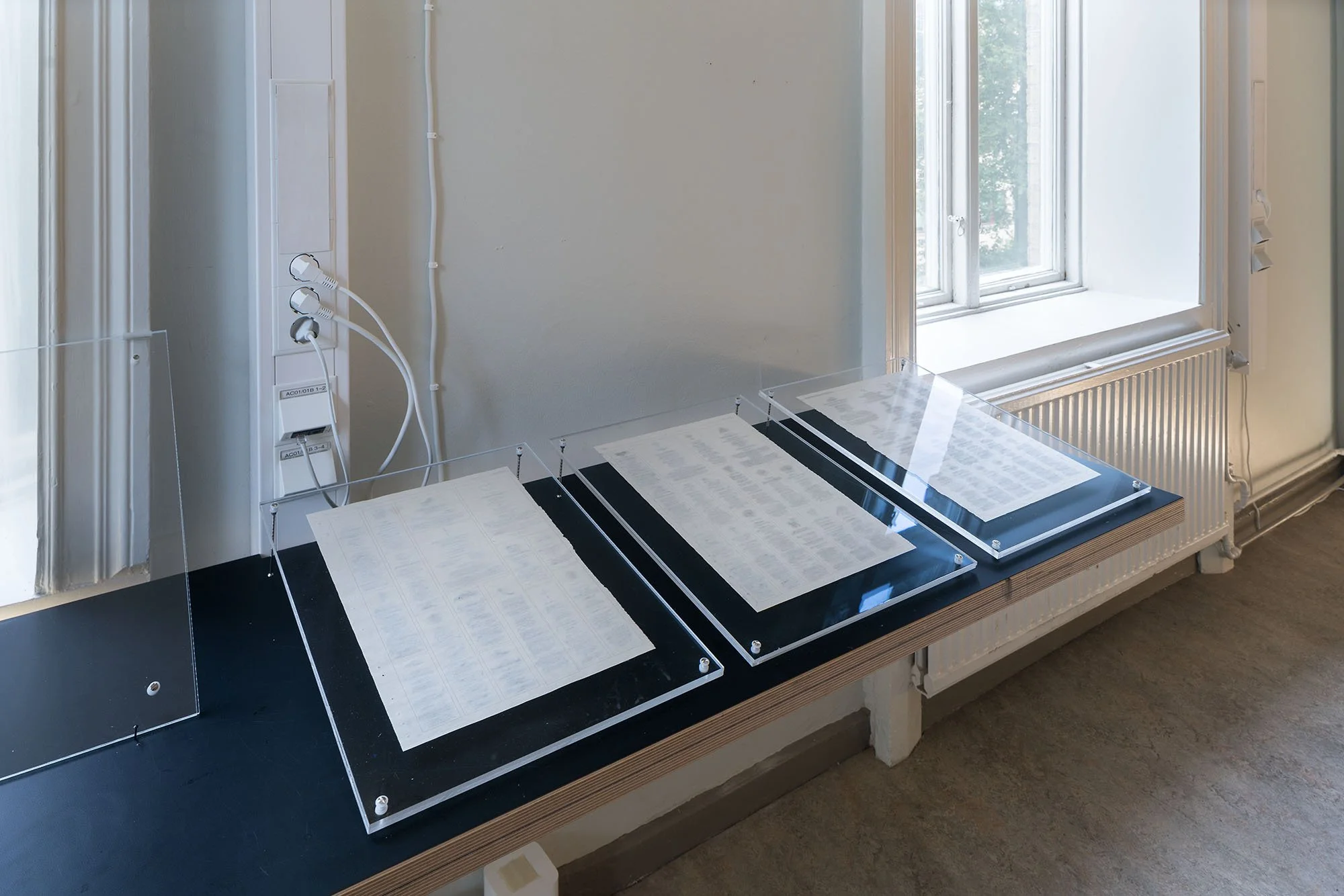 Installation view of three plexi glass panels on a narrow black table top, each displaying a sheet of paper from the Swedish newspaper obituaries.