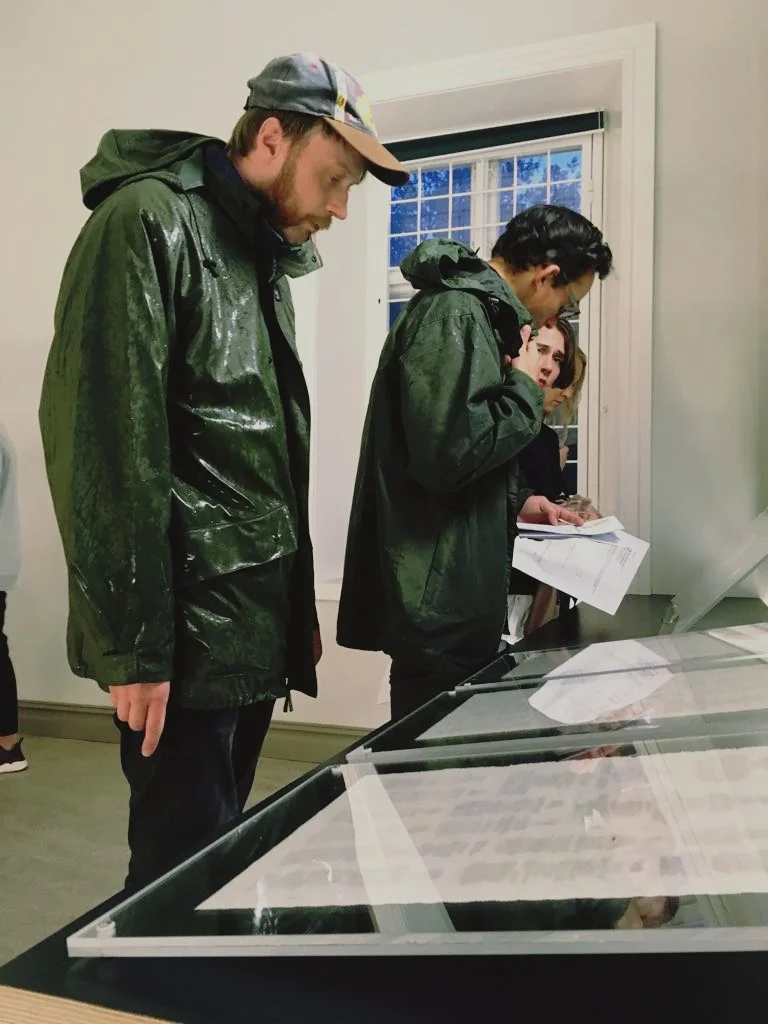 A row of three gallery visitors looking intently at the erased newspaper prints, one of the visitors is wearing a wet dark green raincoat.