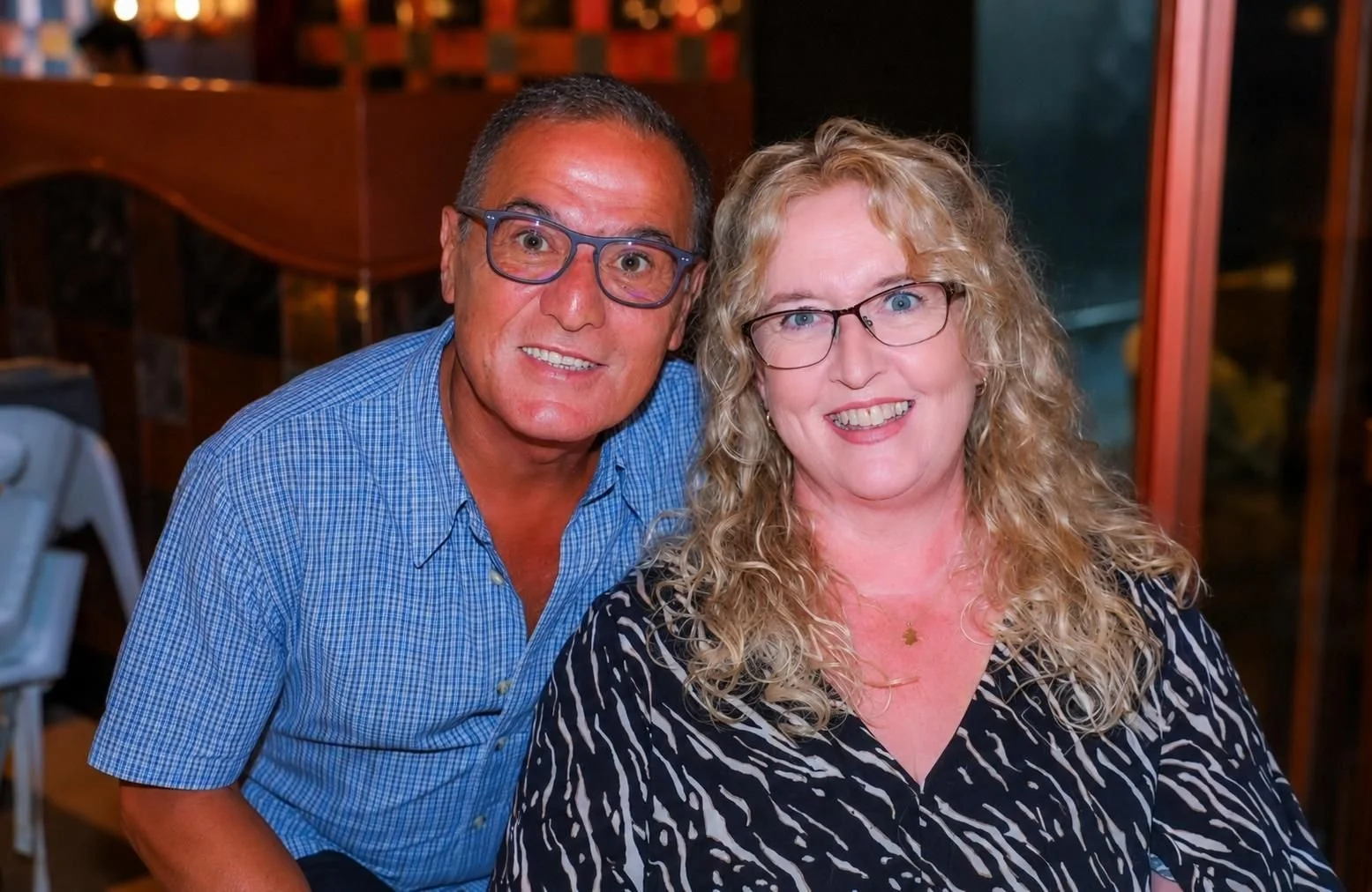 A man and woman smiling, close together, indoors. The man is wearing a blue checked shirt and glasses, the woman has curly blonde hair, glasses, and is wearing a black and white animal print blouse.