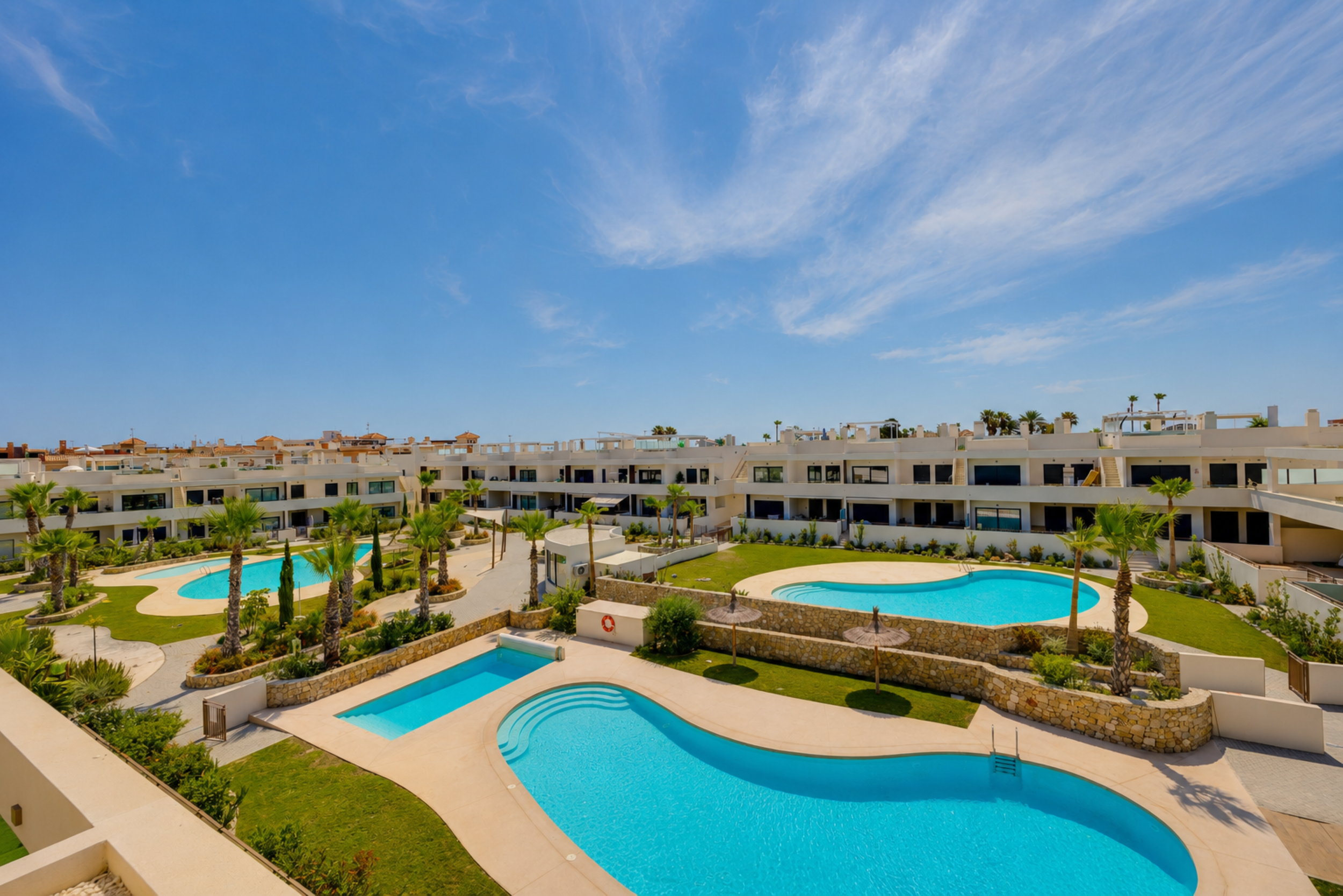 Aerial view of modern white residential buildings with balconies, surrounded by lush greenery, tall palm trees, and a paved courtyard with a stone wall.