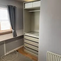 Empty white wardrobe with open space and drawers, in a room with beige walls and a window with blue curtains.