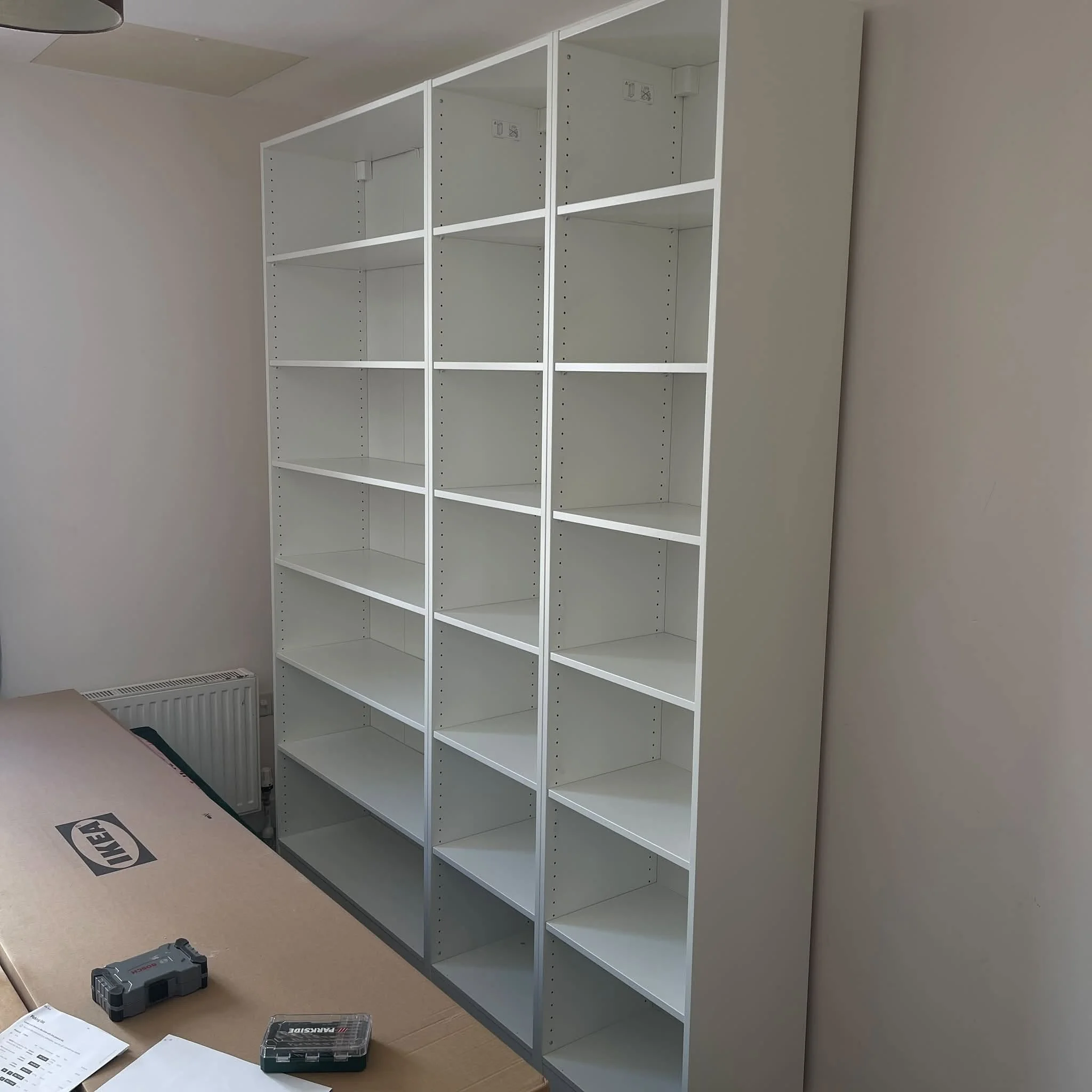 Empty white shelving unit with six compartments in a room with a beige wall. Part of a brown table with tools and papers is visible in the foreground, and an IKEA cardboard box is on the floor.