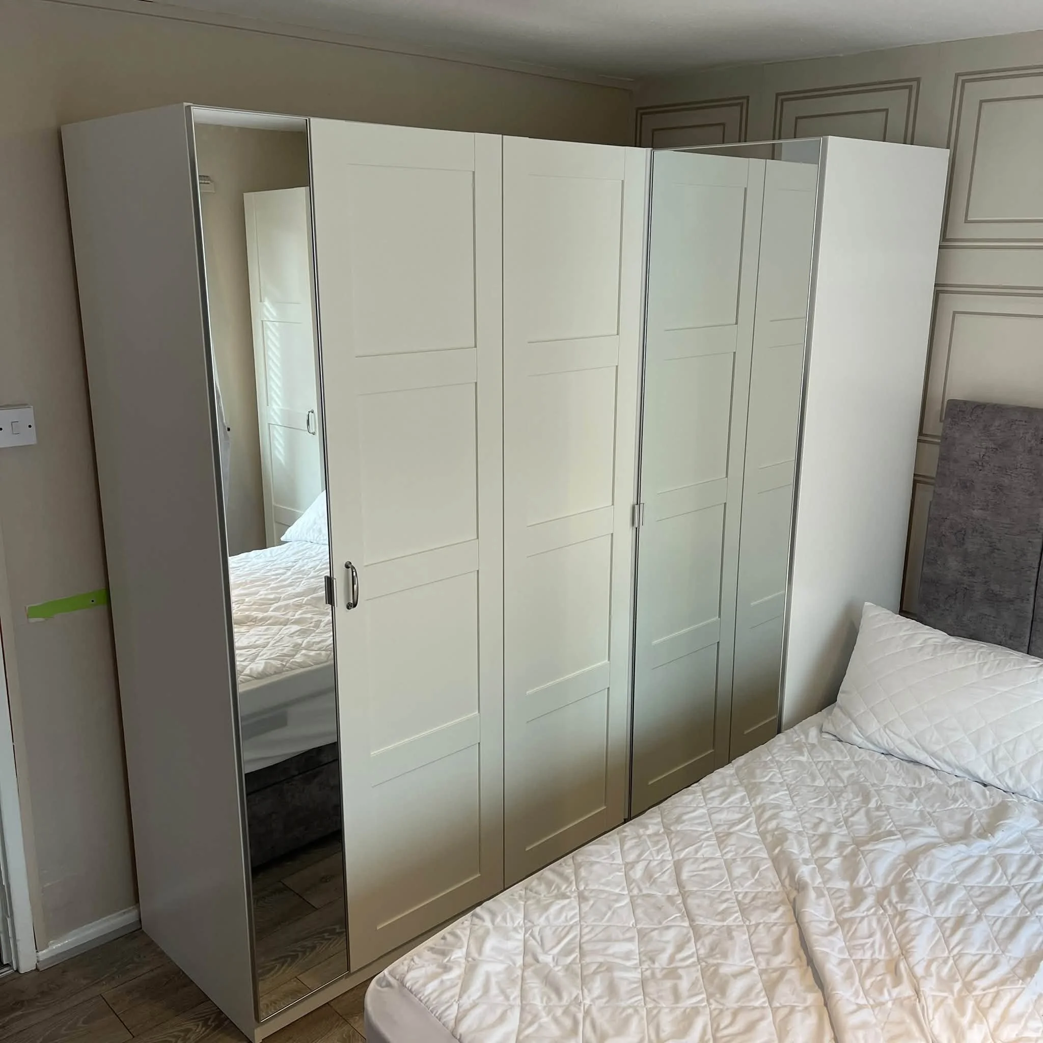 A white wardrobe with mirrored doors in a bedroom, next to a bed with a quilted white blanket.