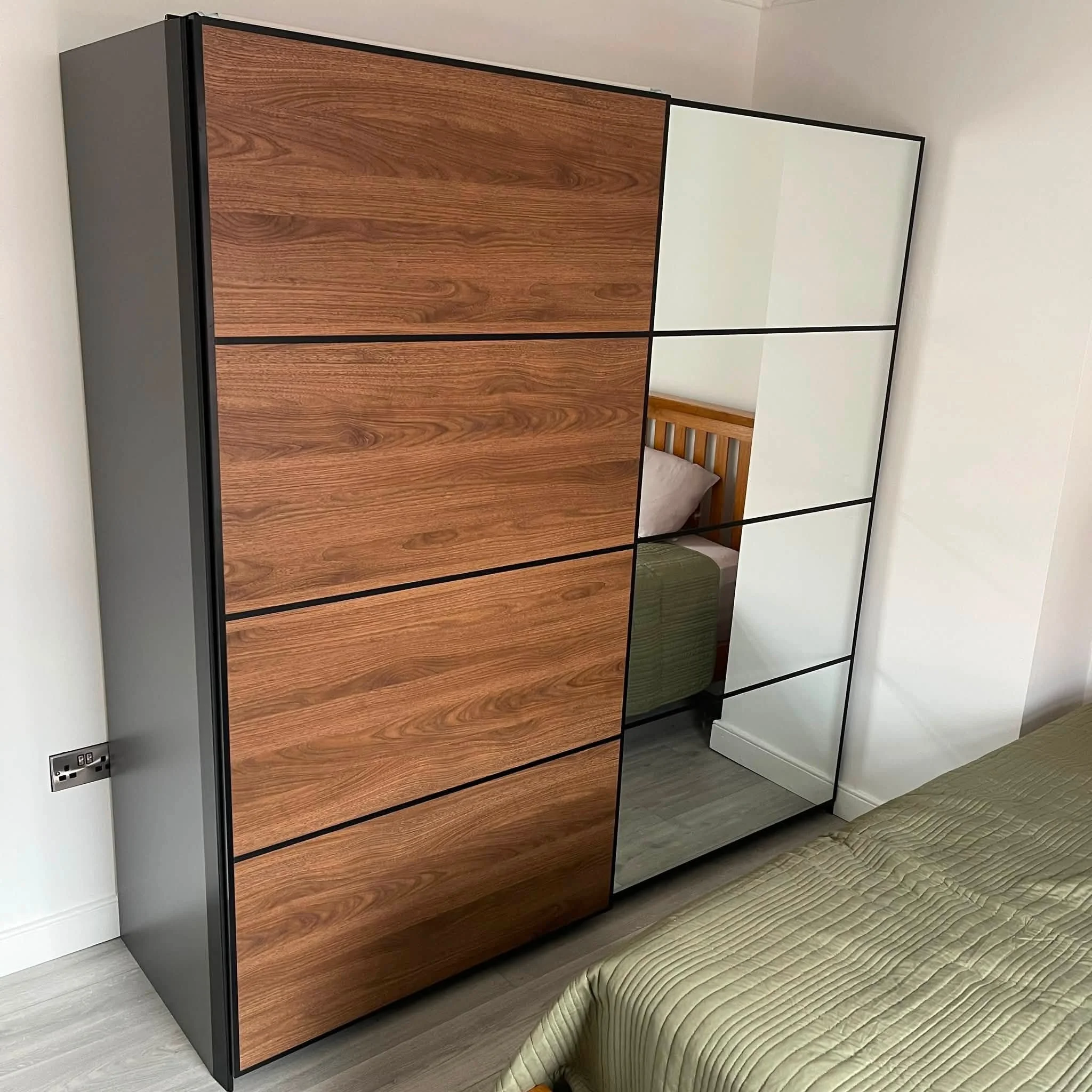 A modern wardrobe with wood and white panels and black lines, placed next to a bed with a beige striped blanket, in a room with white walls and light hardwood floors.