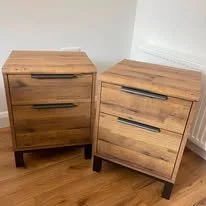 Two wooden nightstands with drawers and black handles, placed on a wooden floor near a white radiator.