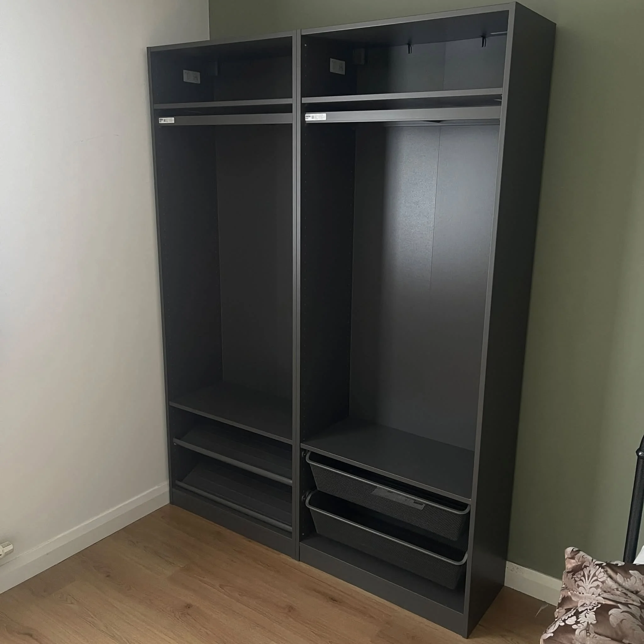 Empty black metal shelving unit with four open shelves, positioned against a light green wall in a room with light wood flooring.