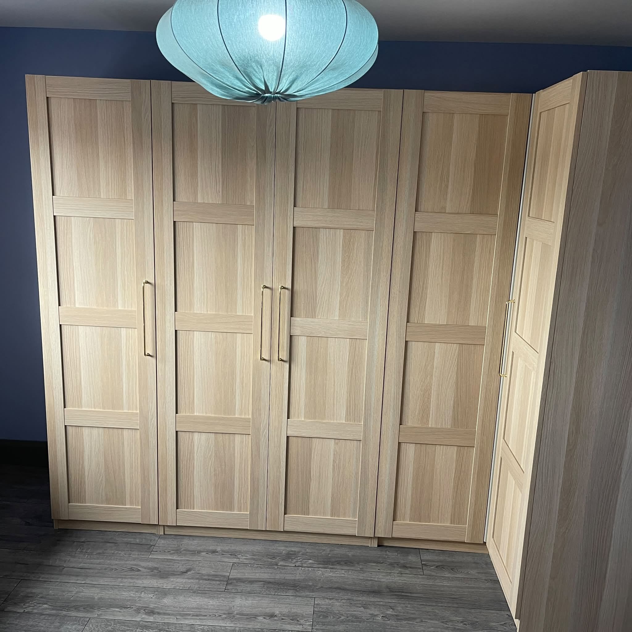 Light wood wardrobe with paneled doors and golden handles, standing against a blue wall in a room with dark wood floor and a blue paper lantern ceiling light.