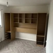 Empty wooden built-in desk with shelves and cabinets in a room with light-colored walls and herringbone-patterned flooring.