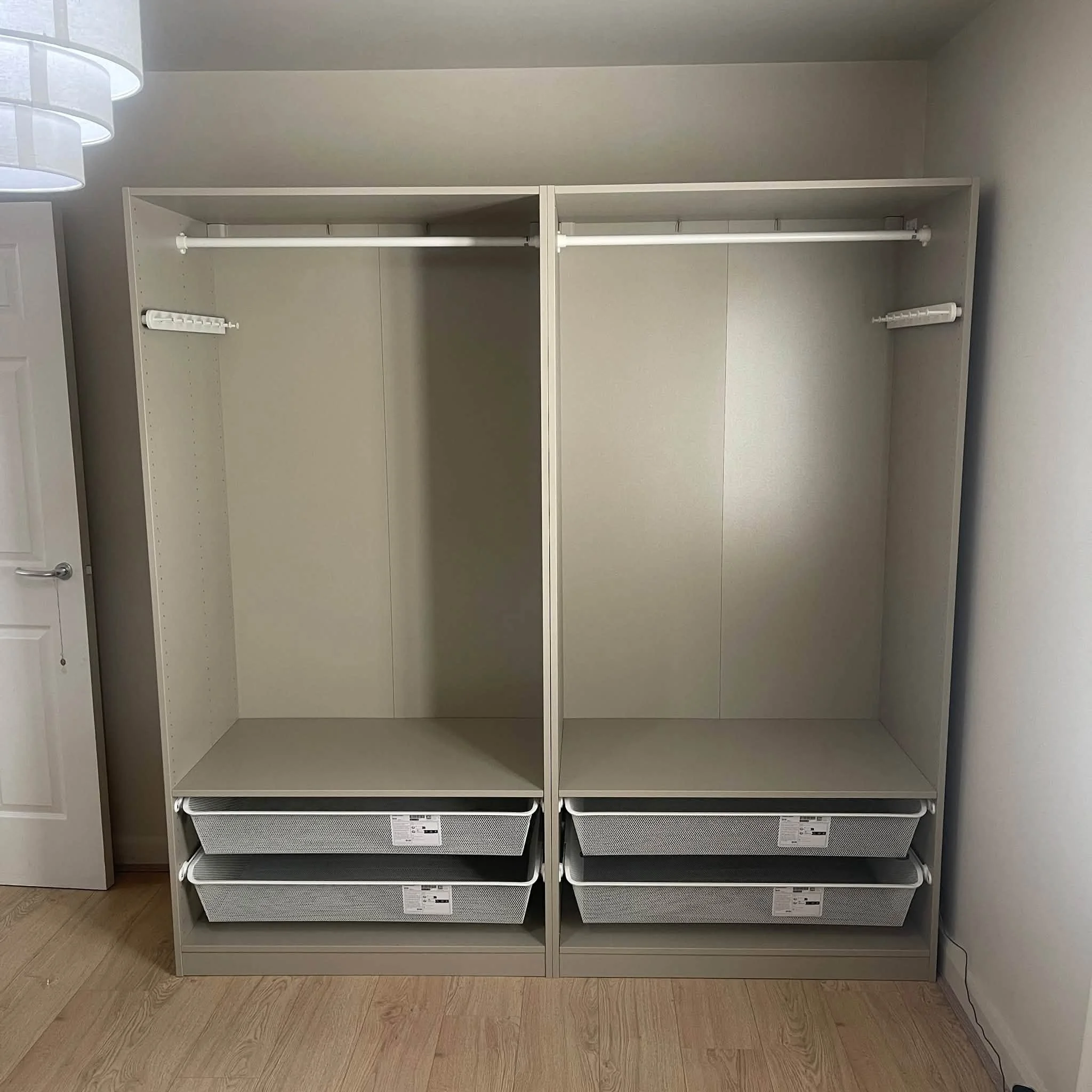 Empty white closet with four mesh baskets on the bottom shelves, two white rods for hanging clothes, and a small hook on the side wall, against a light-colored wall and wooden floor.