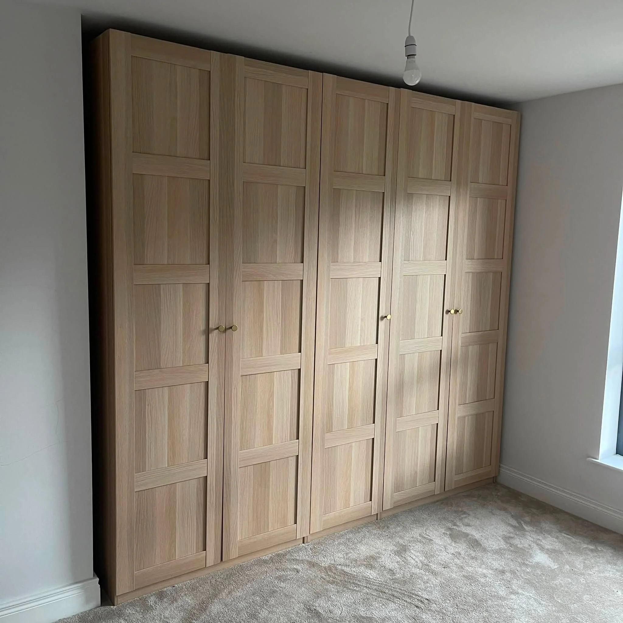 A large wooden wardrobe with multiple doors and round golden knobs, situated in a room with white walls, a beige carpet, and a ceiling light fixture.
