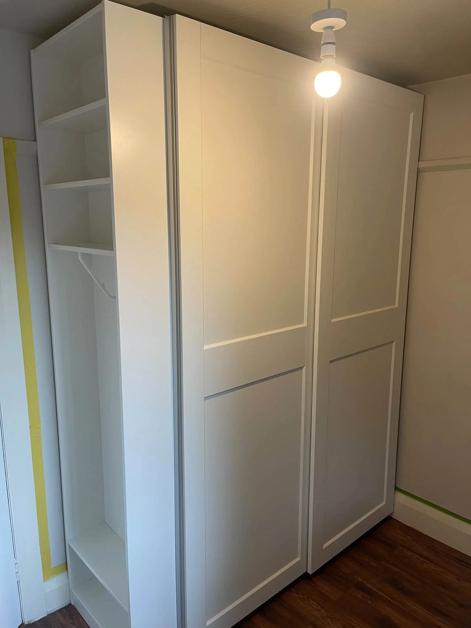 A cream-colored built-in cabinet with open shelving and closed doors, located against a wall with hardwood flooring and a ceiling light bulb.