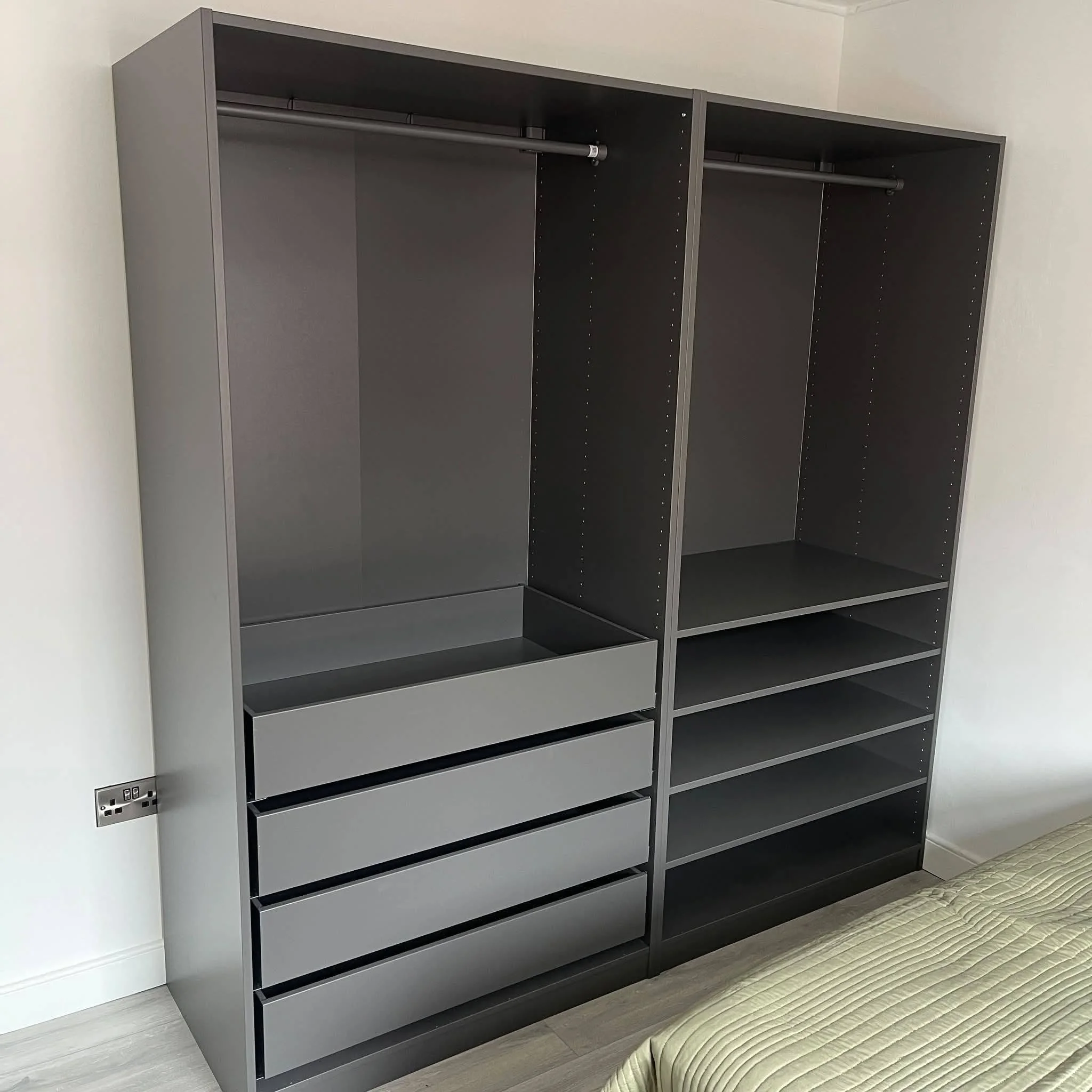 Empty black metal shelving unit with multiple sections, positioned next to a wall and near a bed with a green and beige striped bedspread.
