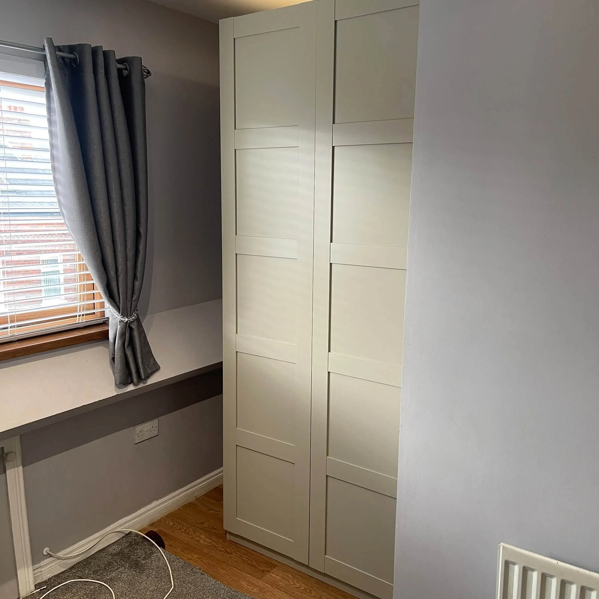 A white wardrobe or closet unit with multiple drawers and cabinet doors is positioned against a wall in a room. To the left, there is a window with closed blinds and dark grey curtains. Below the window, an electrical outlet is visible, with a white 