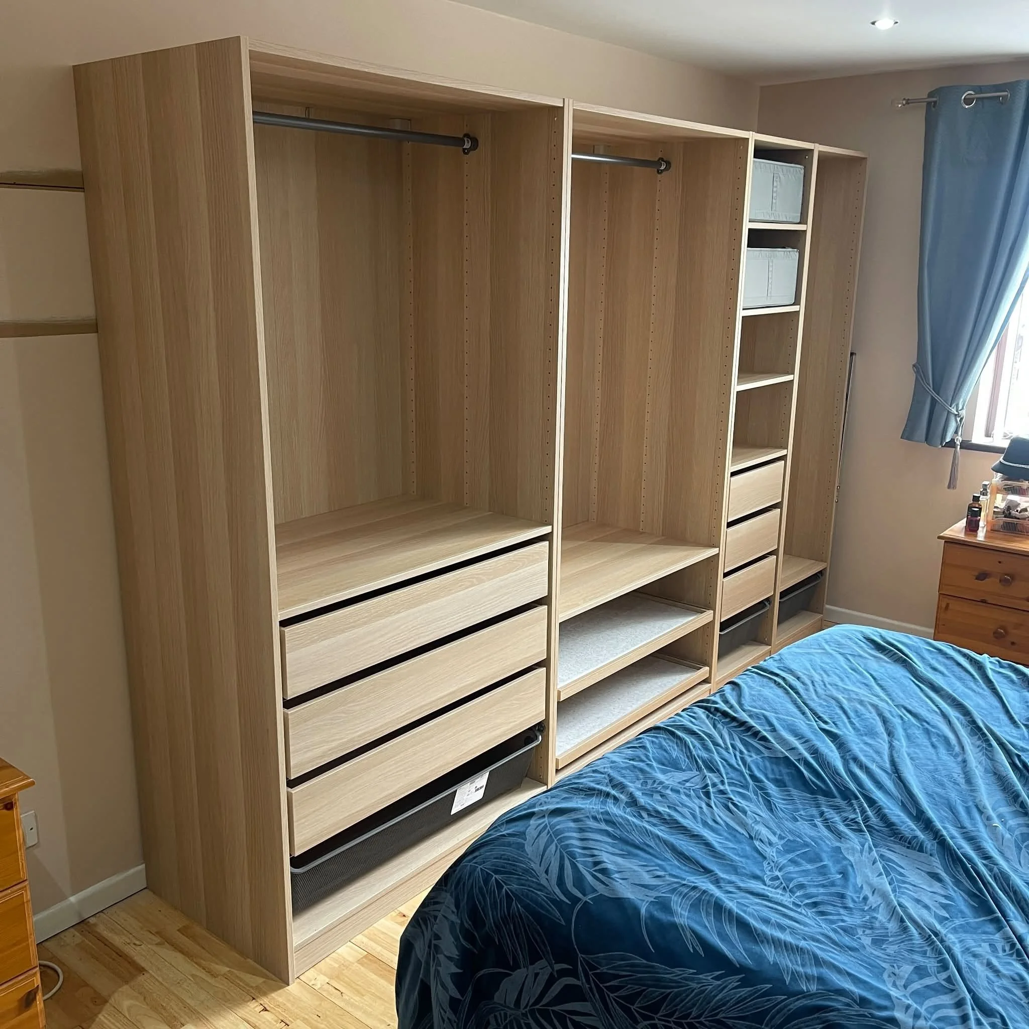 Unfinished wooden closet frame in a bedroom, with a bed with a blue patterned comforter in the foreground and a window with a blue curtain on the right side.