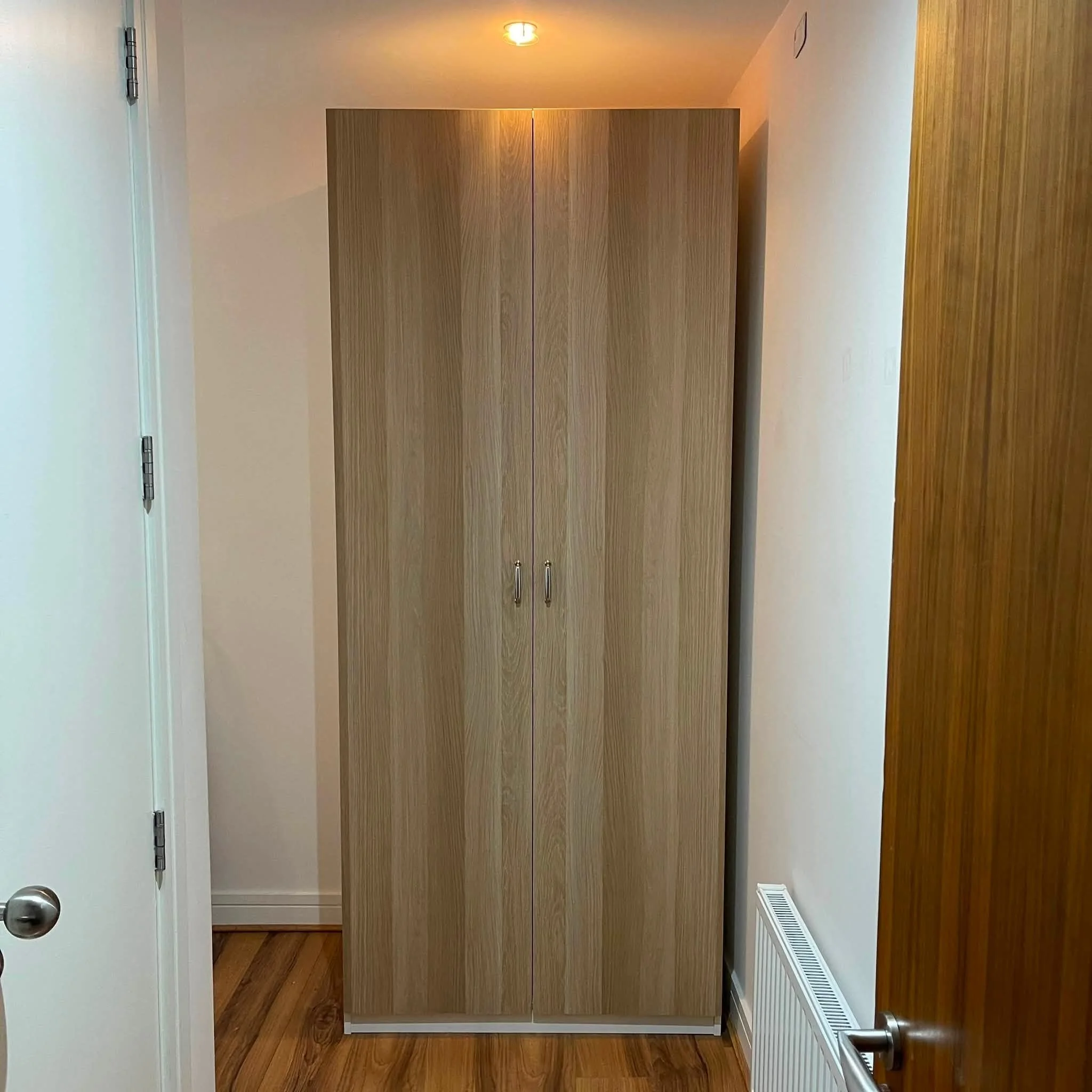 Wooden cabinet in a narrow hallway with white walls, a ceiling light, and a radiator on the right side.