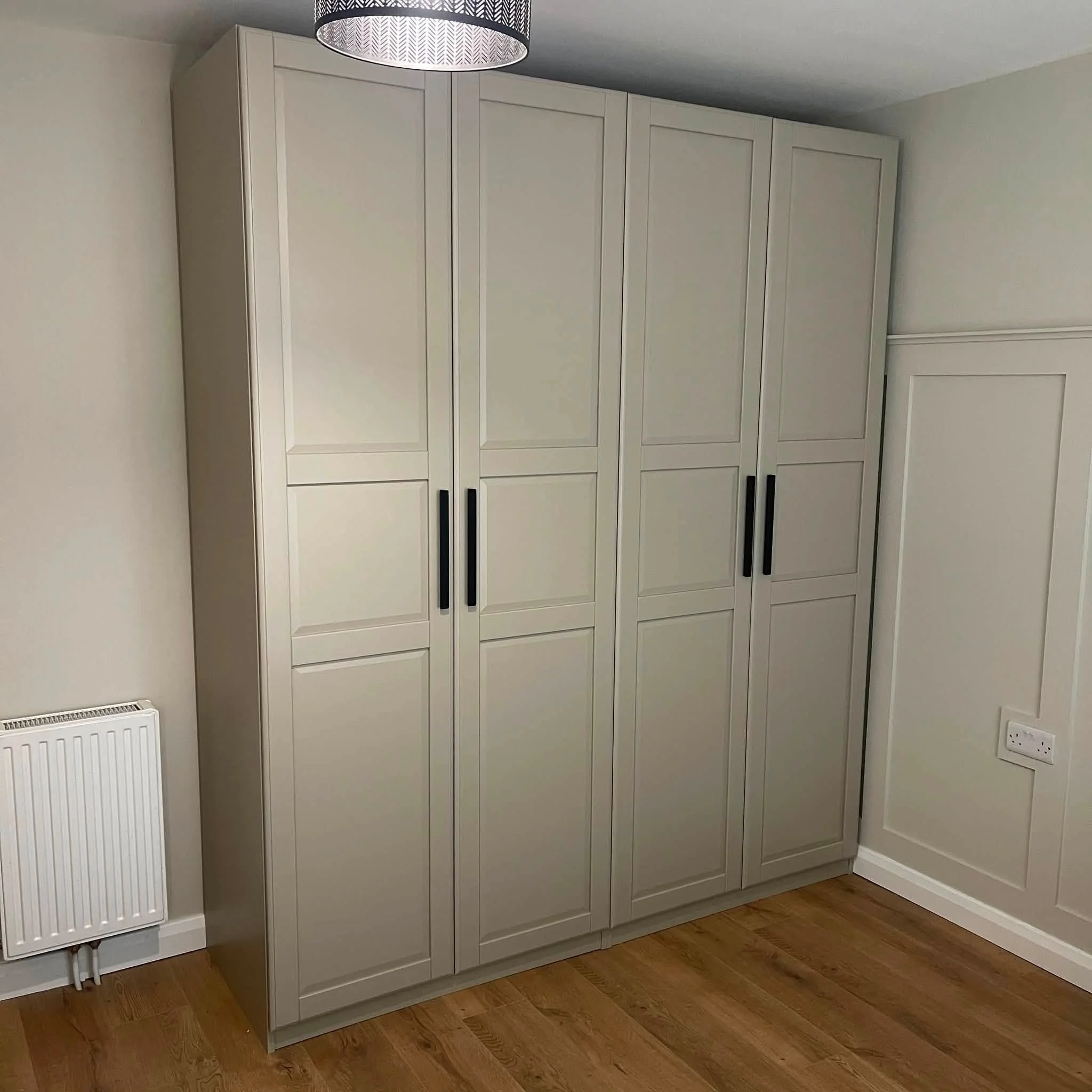 A row of cream-colored built-in cabinets with black handles in a room with wooden flooring. There is a white radiator with pipes on the left side and a white electrical socket on the right side near the floor. The ceiling has a patterned light fixtur