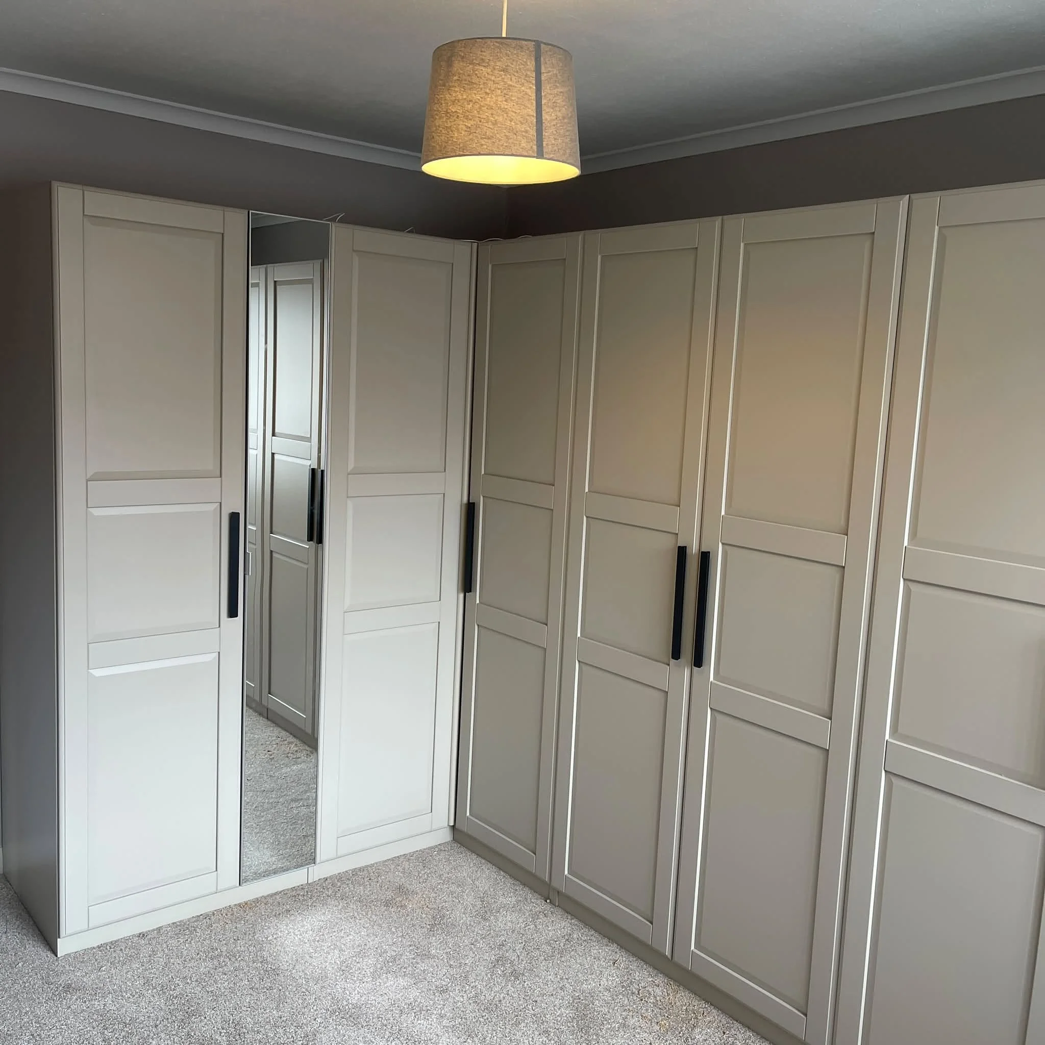 A corner of a room with white built-in cabinets, a mirror, and a ceiling light fixture with a fabric shade, beige carpeted floor, and light gray walls.