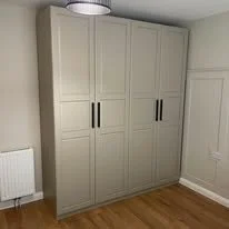 A large beige wardrobe with multiple doors and black handles, positioned against a light-colored wall with a wooden floor.