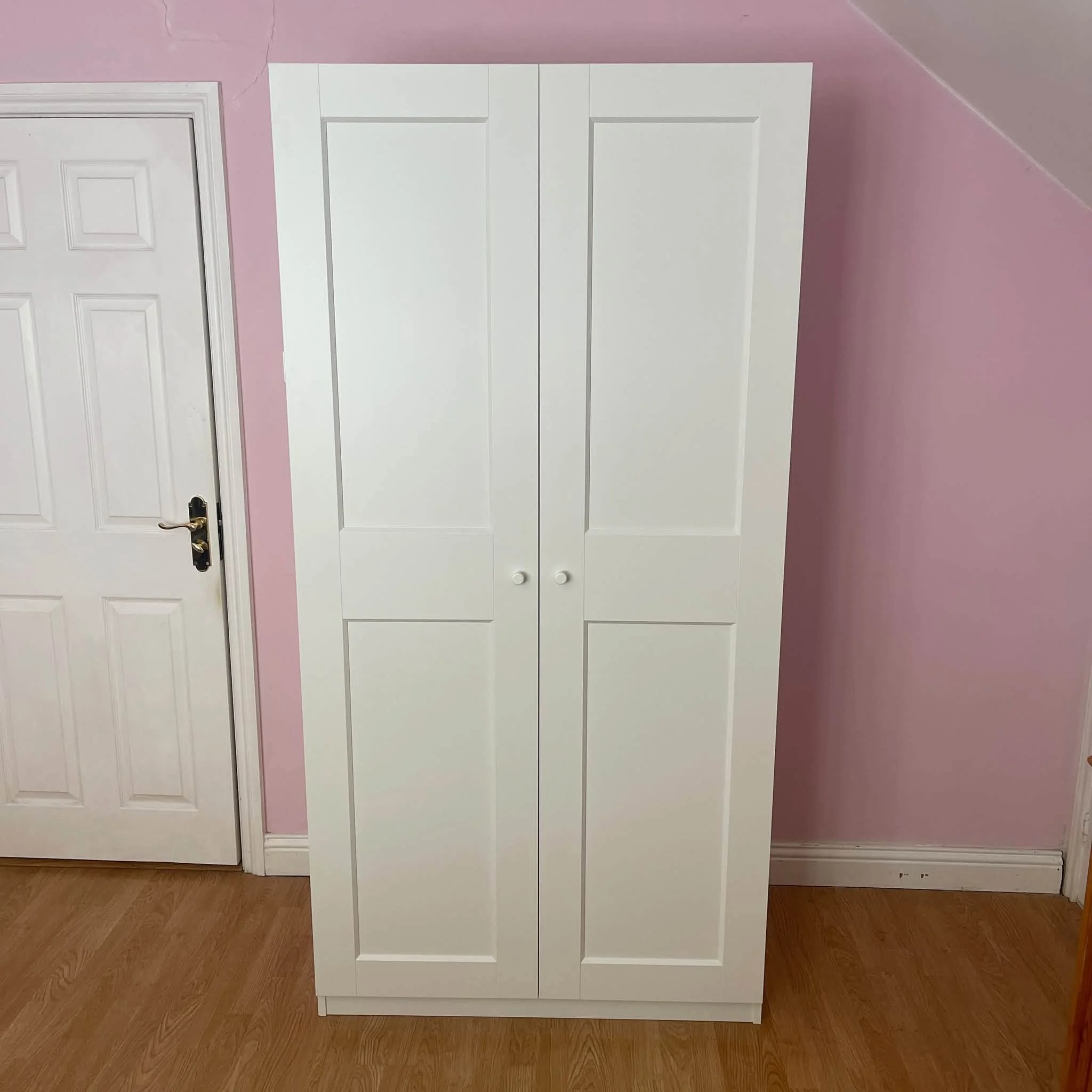 White cabinet with two doors and knobs, standing against a pink wall with corner moldings, on a wooden floor.