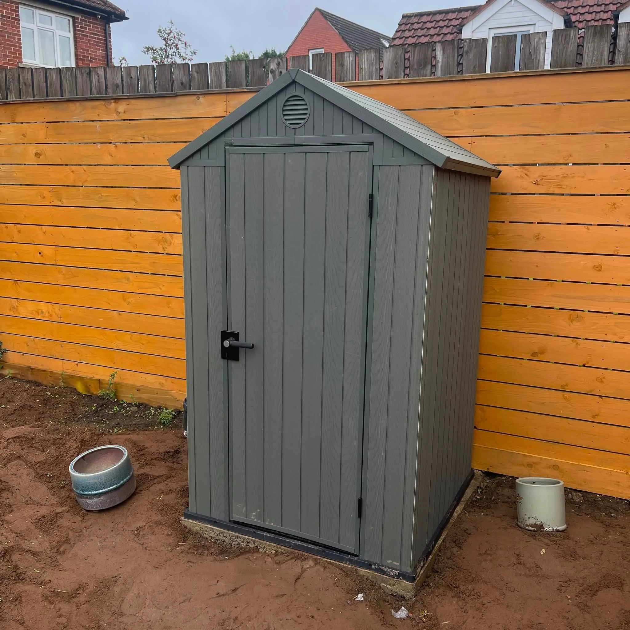 A small grey metal shed with a pitched roof, located in a backyard with a wooden fence. The ground is bare dirt, and there are two metal buckets on the ground nearby.