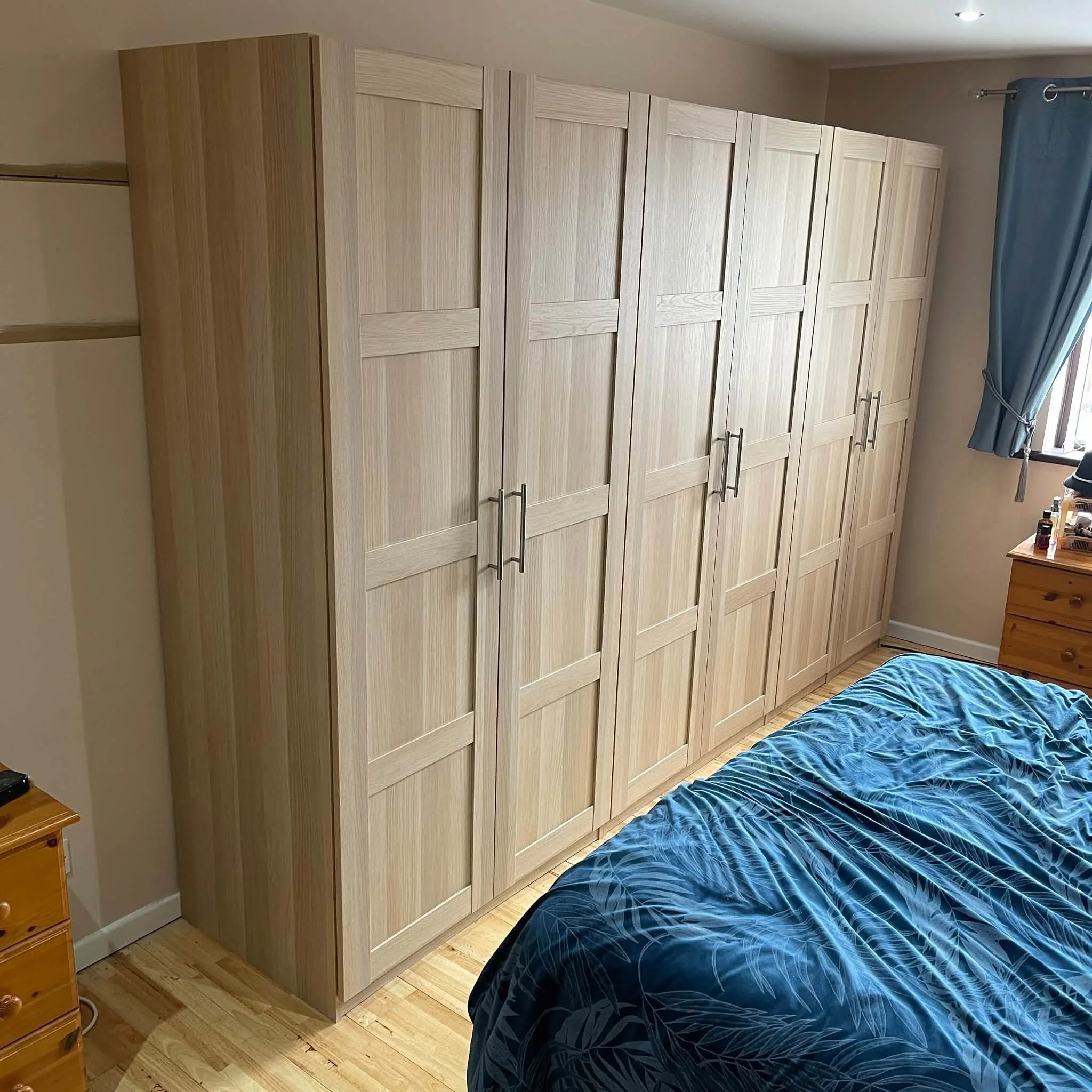 Light wood wardrobe with multiple doors and silver handles in a bedroom, next to a blue bedsheet with a leafy pattern, a wooden nightstand, and a window with blue curtains.