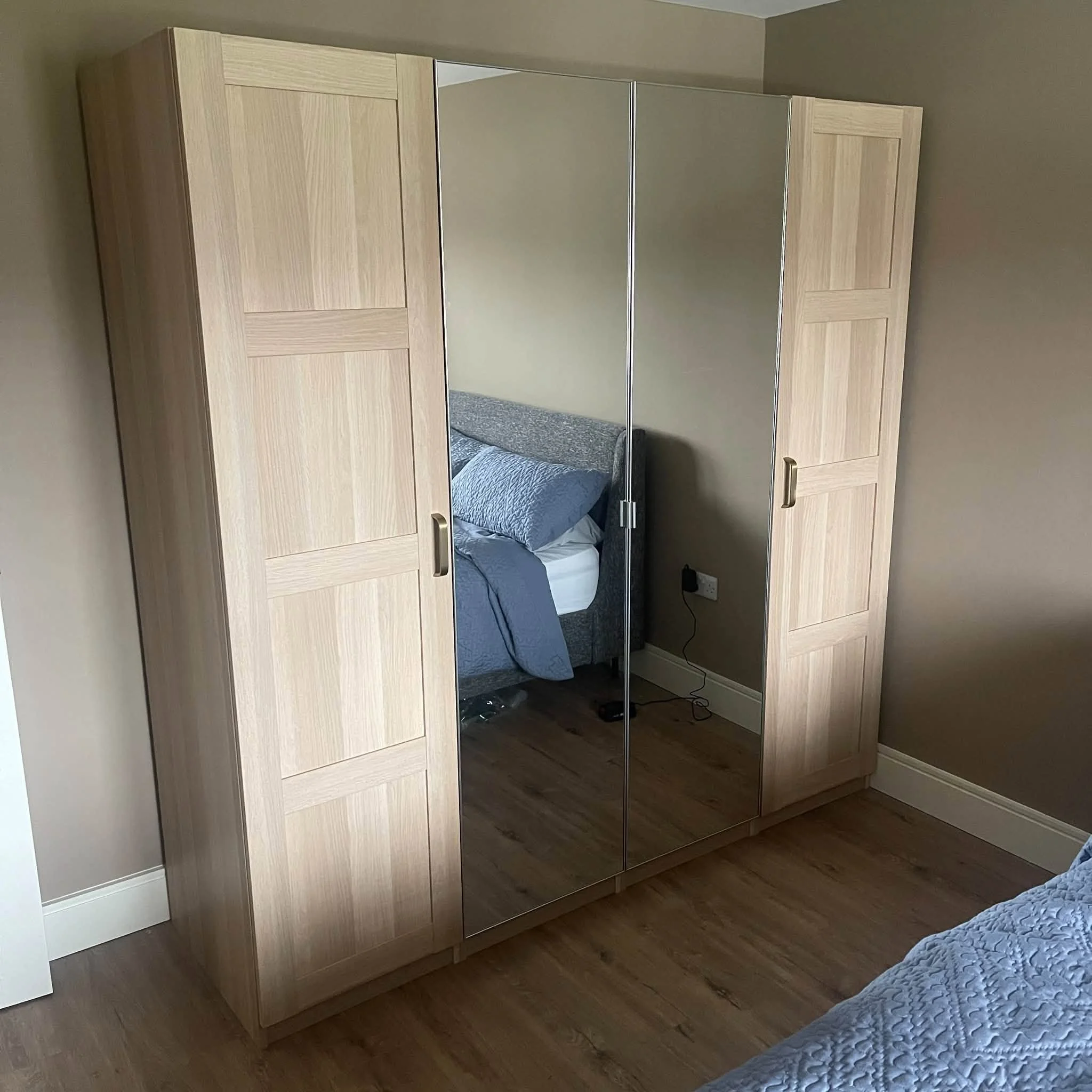 A light wood wardrobe with sliding mirror doors in a room with gray walls and wooden flooring, partially reflecting a bed with a gray quilt.