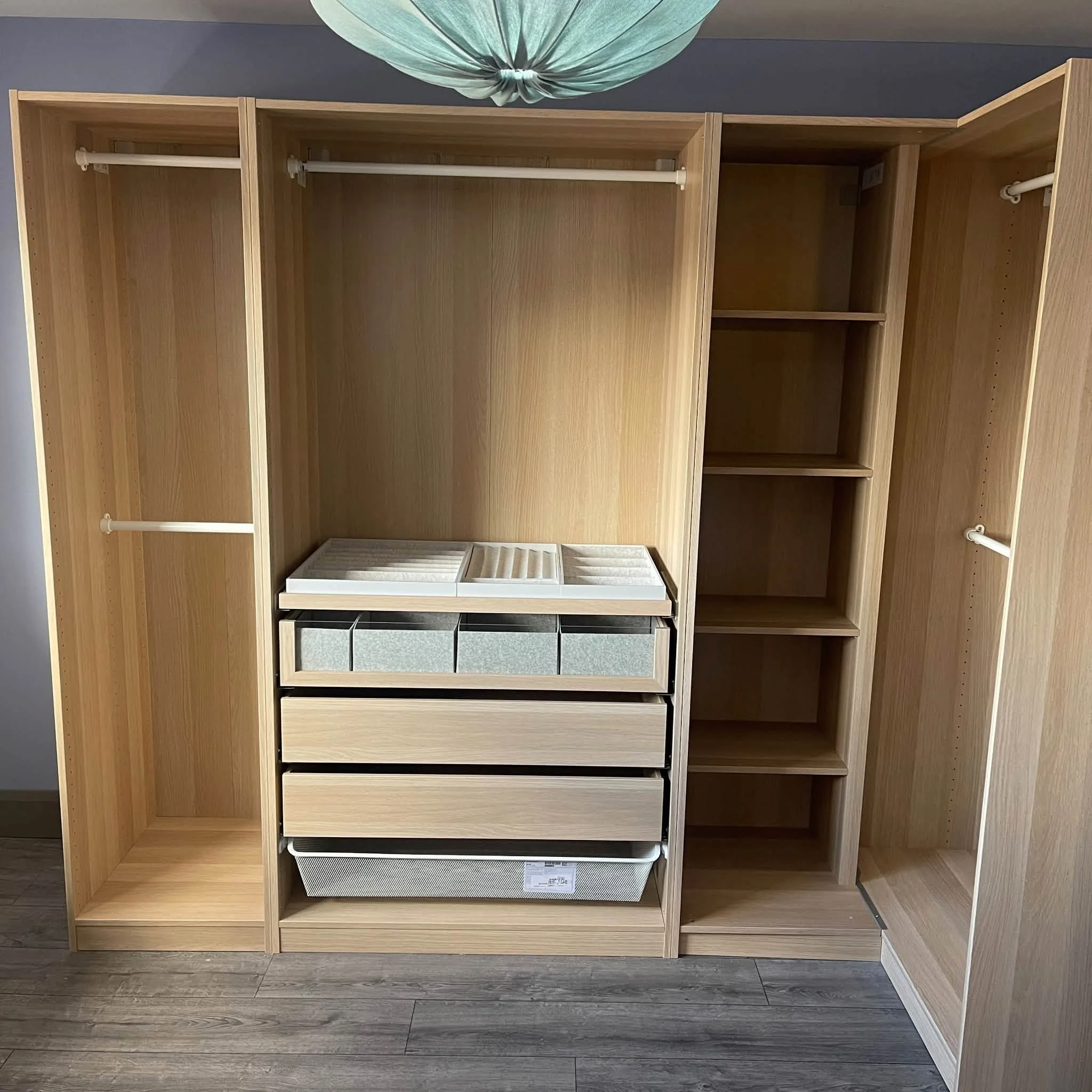Empty wooden closet with multiple compartments and rods for hanging clothes, located against a gray wall with wood flooring.