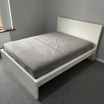 White bed frame with gray mattress in a room with gray walls and carpeted floor.