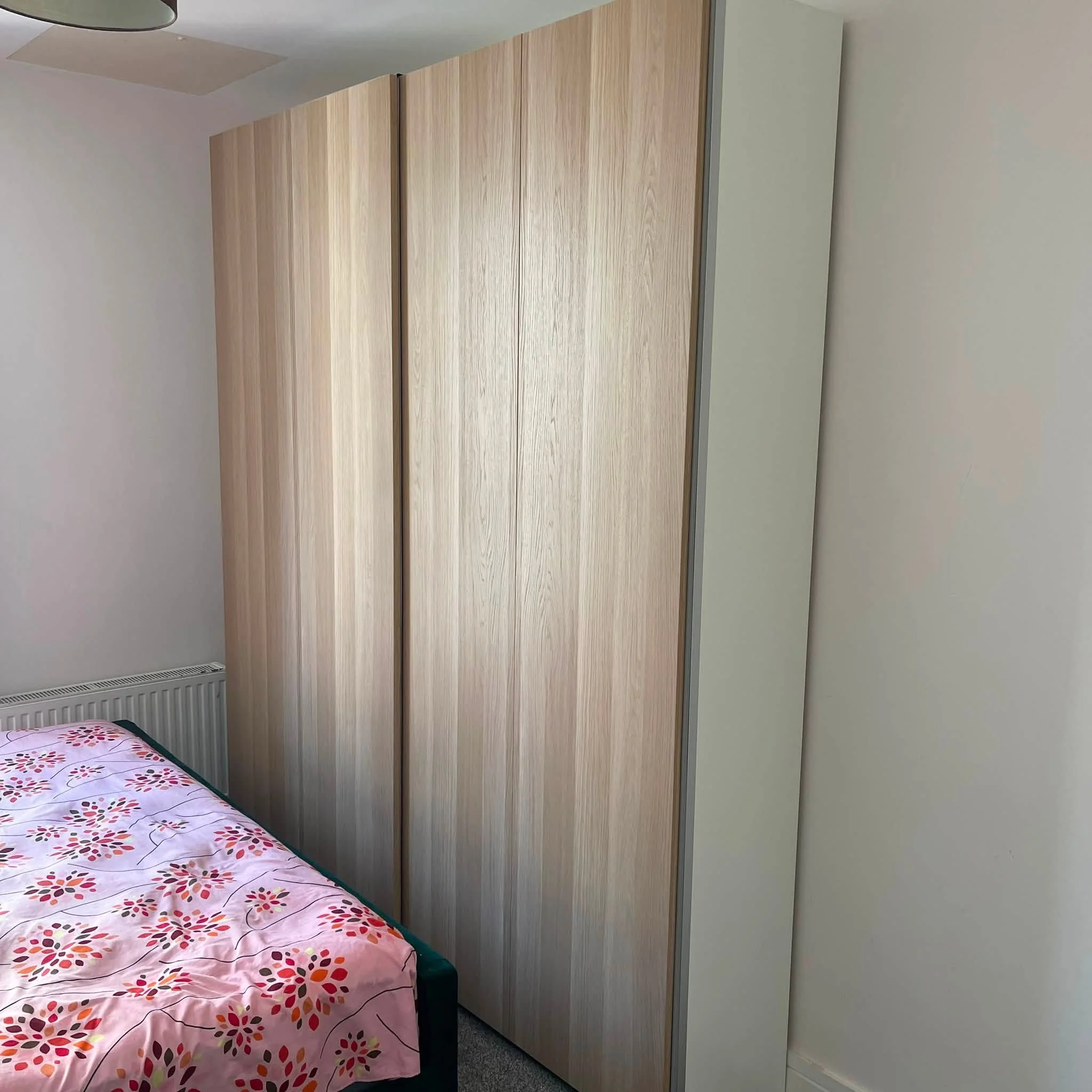 Bedroom with a bed featuring pink floral bedding, a gray carpet, and a tall wooden wardrobe against the wall.