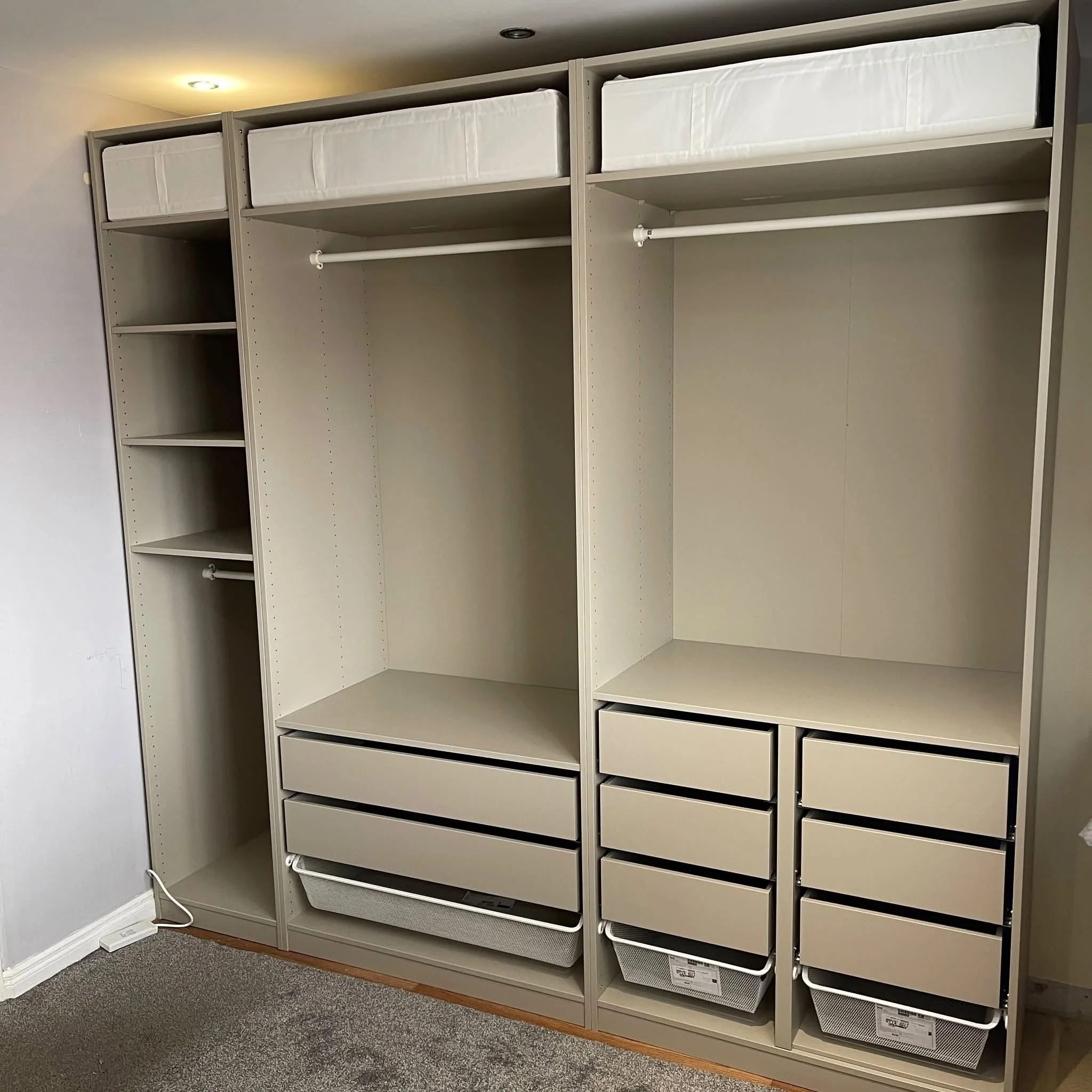 Empty beige shelving unit with drawers, shelves, and storage baskets, situated next to a white wall and gray carpet in a room with ceiling lights.