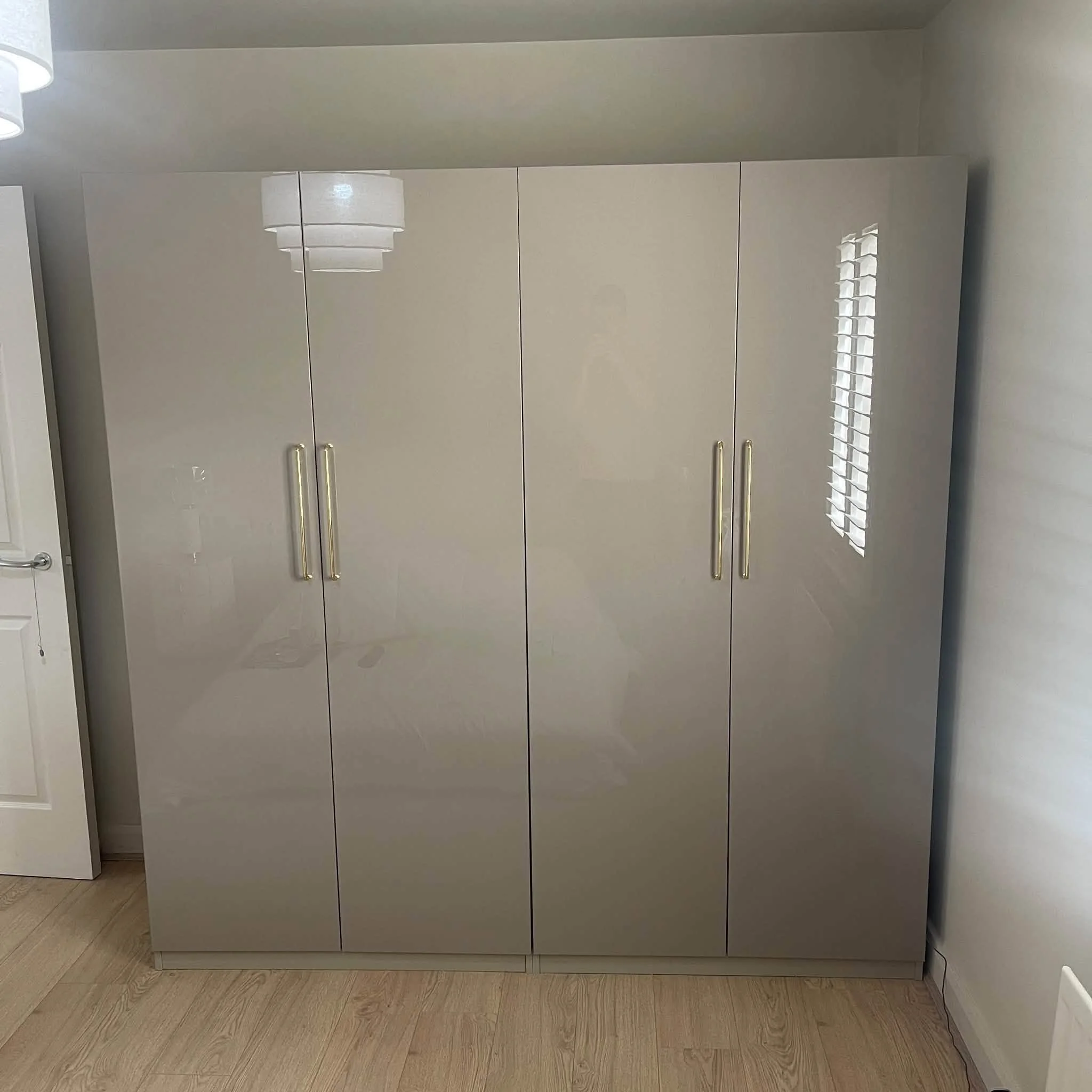 A modern beige kitchen cabinet with gold handles reflected in a mirror, against a white wall with blinds and a floor with wooden planks.