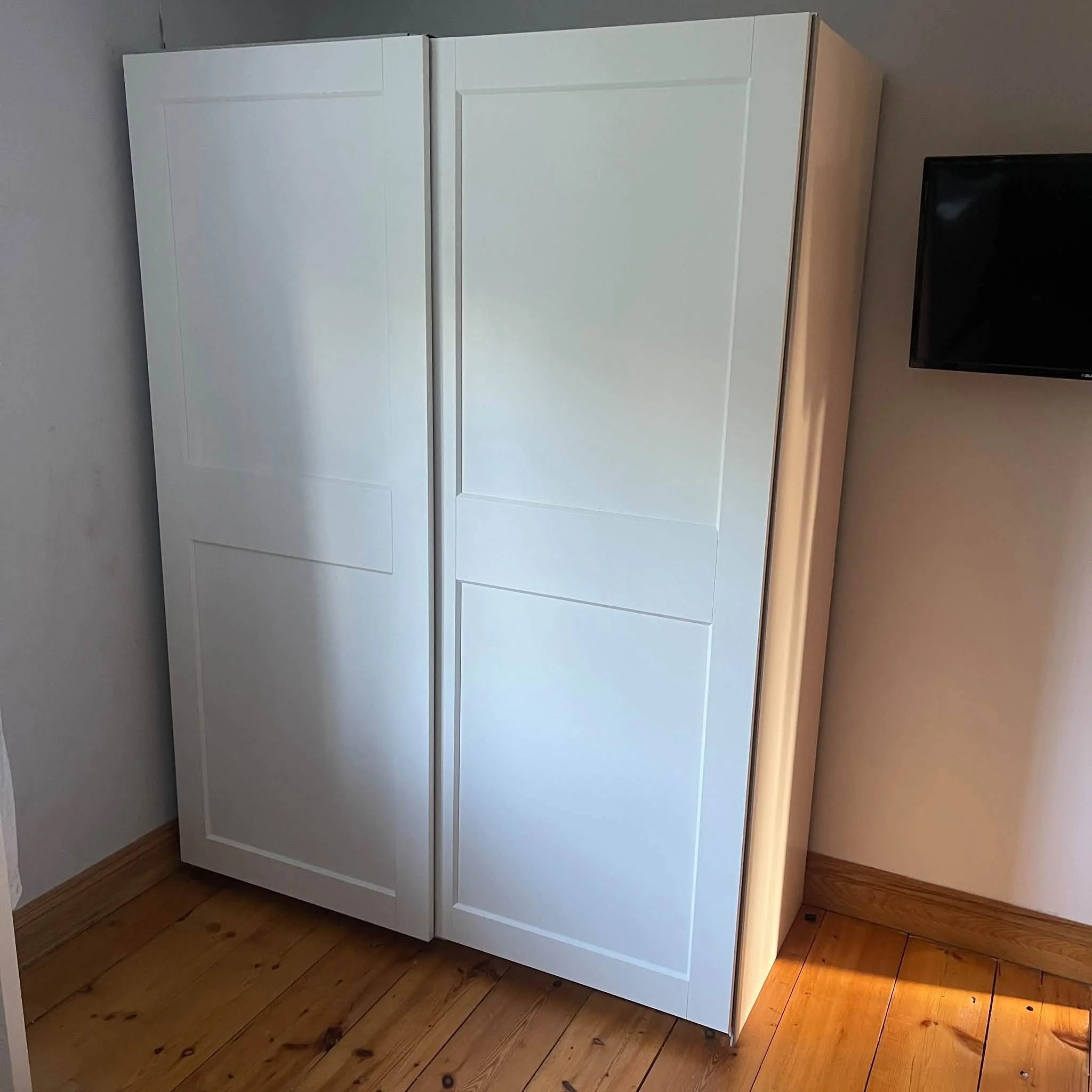 White wardrobe with three doors, placed on a wooden floor next to a wall and a television.