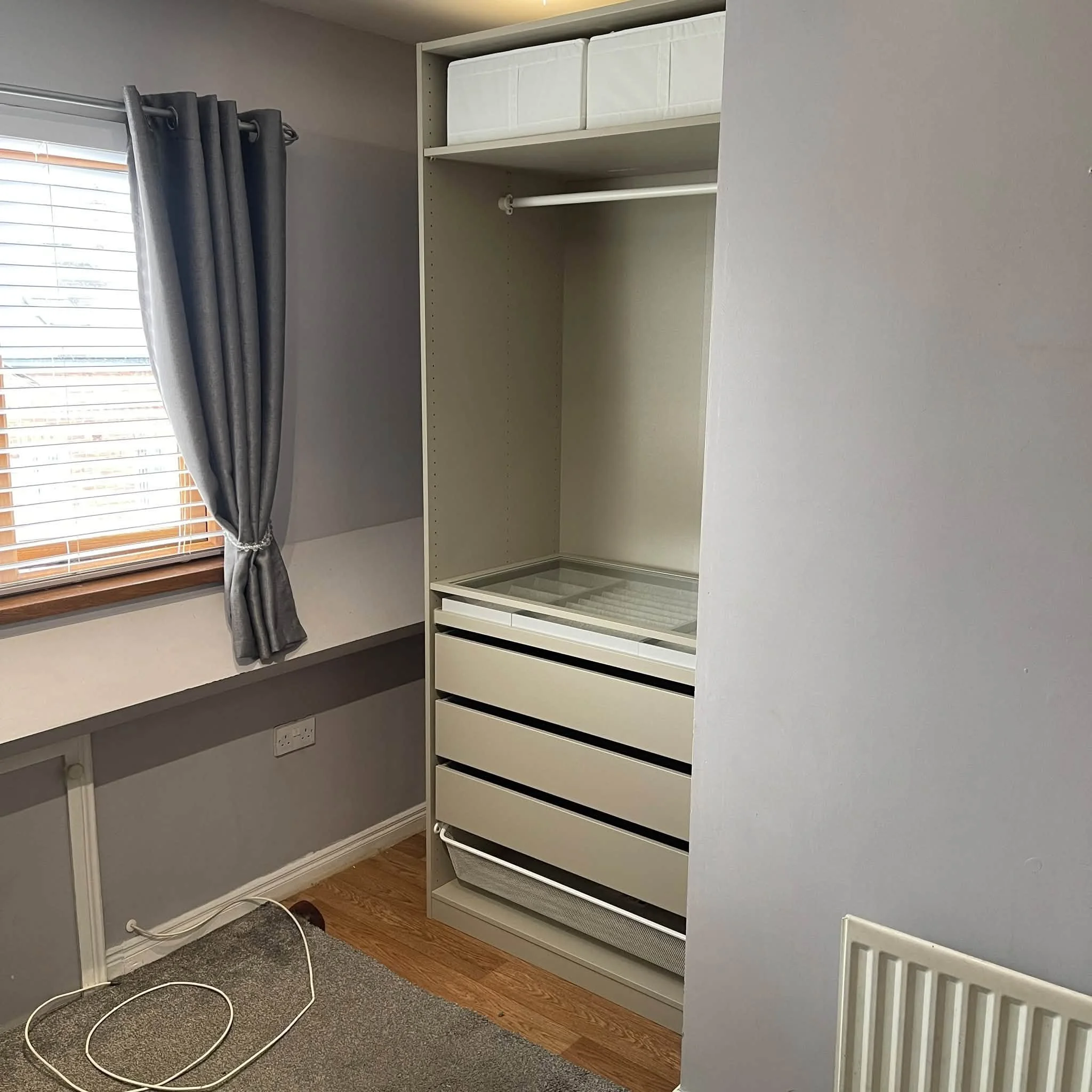 Empty white shelving unit with drawers and a rod, situated next to a window with gray curtains, in a room with wood flooring and gray carpet.