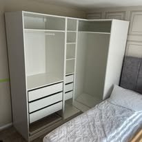 White wardrobe with open doors showing shelves and drawers, beside a bed in a bedroom.