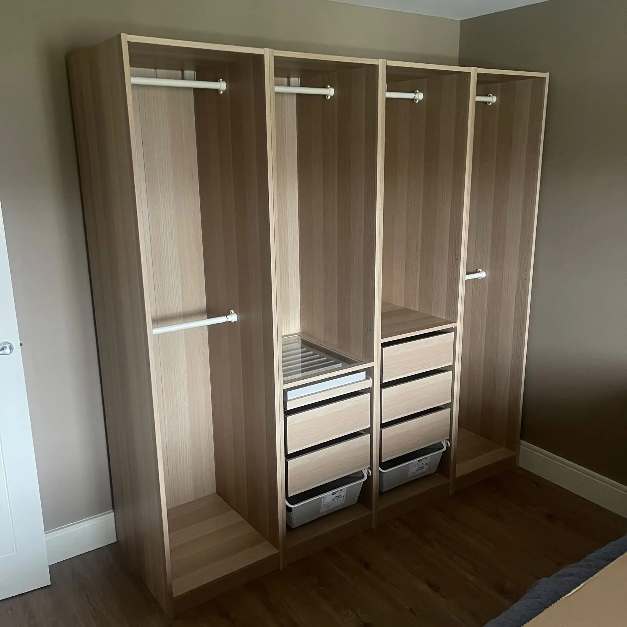 Empty wooden wardrobe with multiple sections and black, white, and brown drawers at the bottom, placed against a beige wall in a room with wooden flooring.