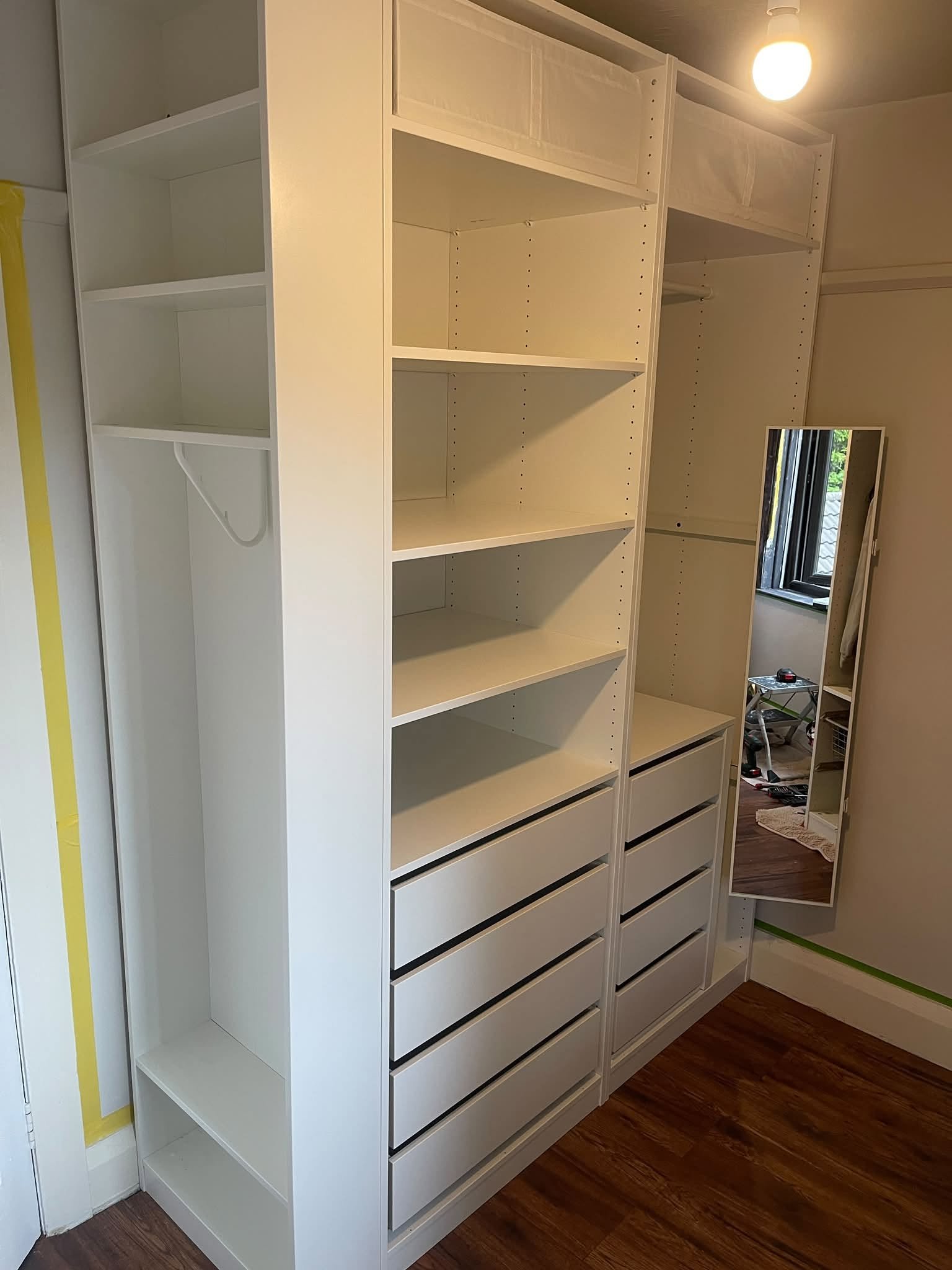 A white built-in shelving unit with multiple empty shelves and drawers, positioned next to a mirror on a wall in a room with wooden flooring and an open window.
