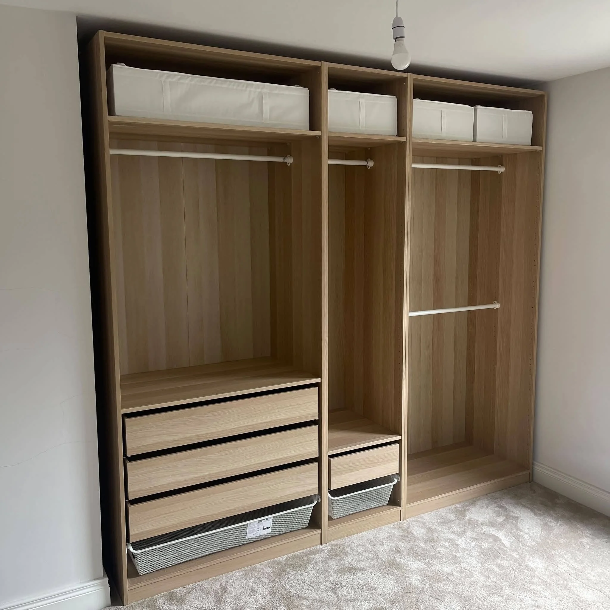 New wooden bedroom wardrobe with open shelves and hanging rods, placed on beige carpeted floor in room with white walls and a ceiling light bulb.