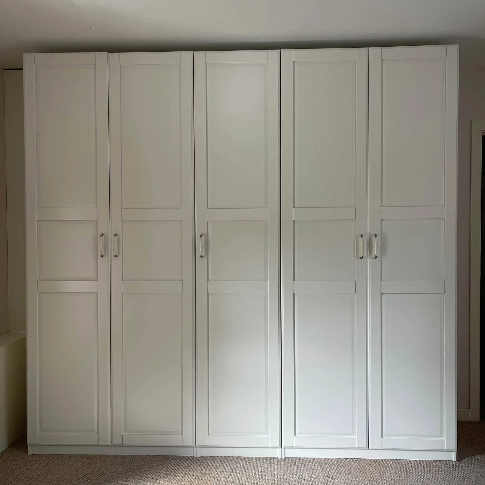 White built-in closet with multiple doors and small silver handles, set against a beige wall and carpeted floor.