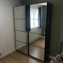 A mirrored wardrobe with sliding doors in a bedroom with white walls, wooden flooring, and a window with blue curtains.