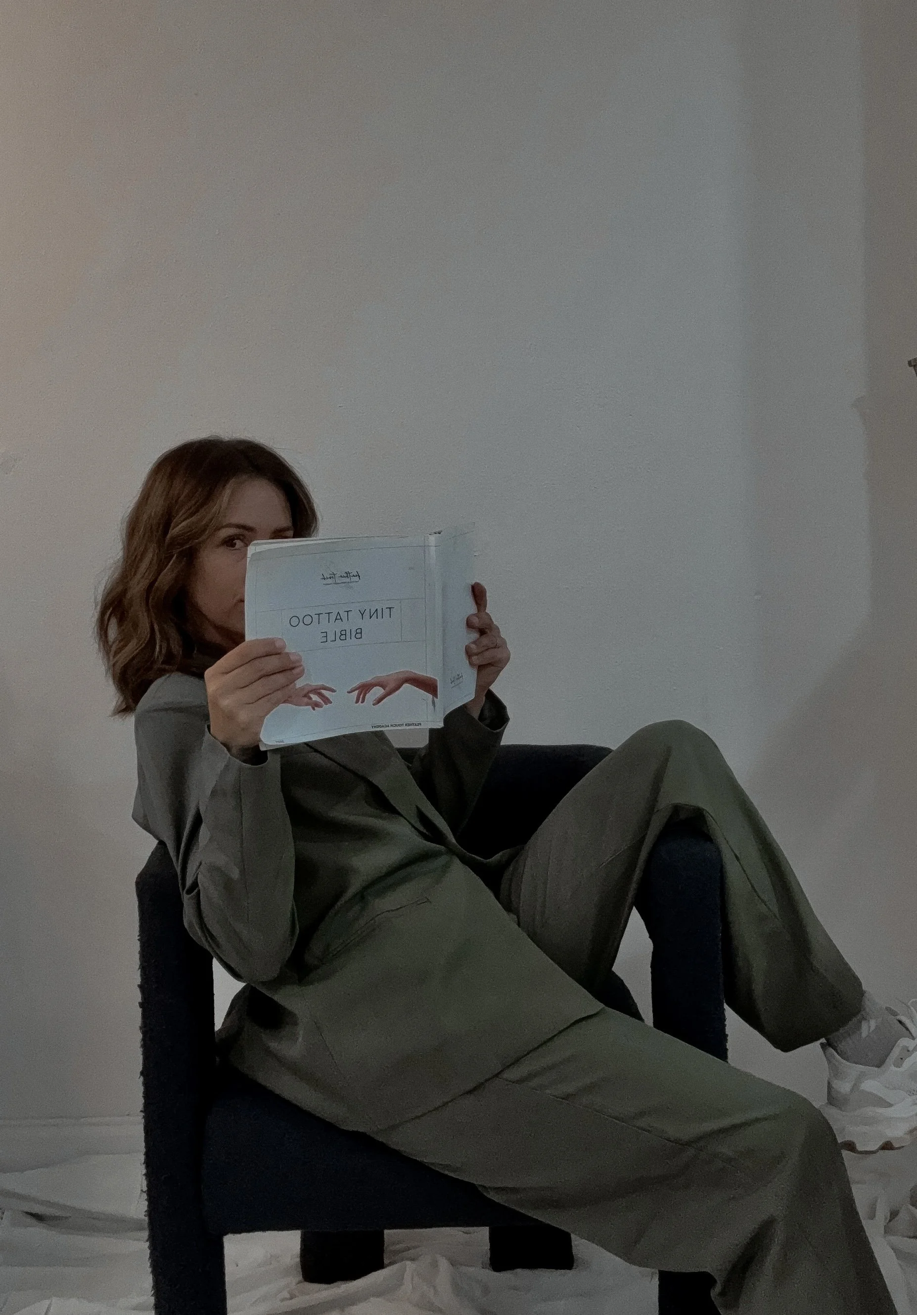 A woman with wavy brown hair holding a book titled "Tiny Tattoo Bible" and placing it in front of her face, sitting on a black chair, with her legs stretched out on a table, in a room with plain white walls and a paper-covered floor.