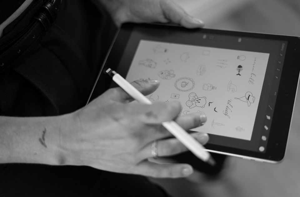 A person holding a stylus and using a tablet with doodles and handwritten notes on the screen in black and white.