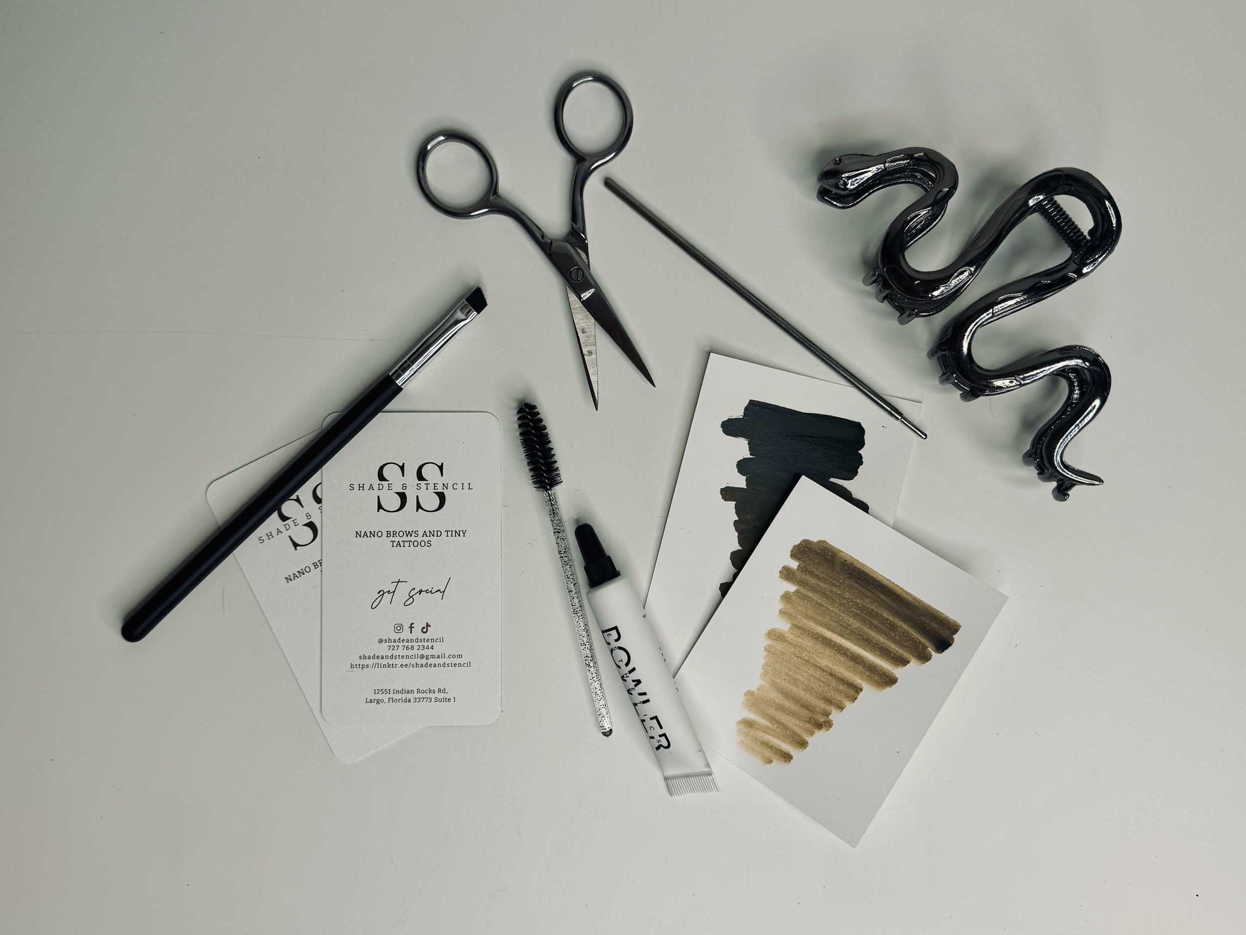 Flat lay of black scissors, a pen, a metal stick, a spool of string, black mascara, and sample swatches of gold, beige, and black paint on a white surface, with business cards for shade and stencil.