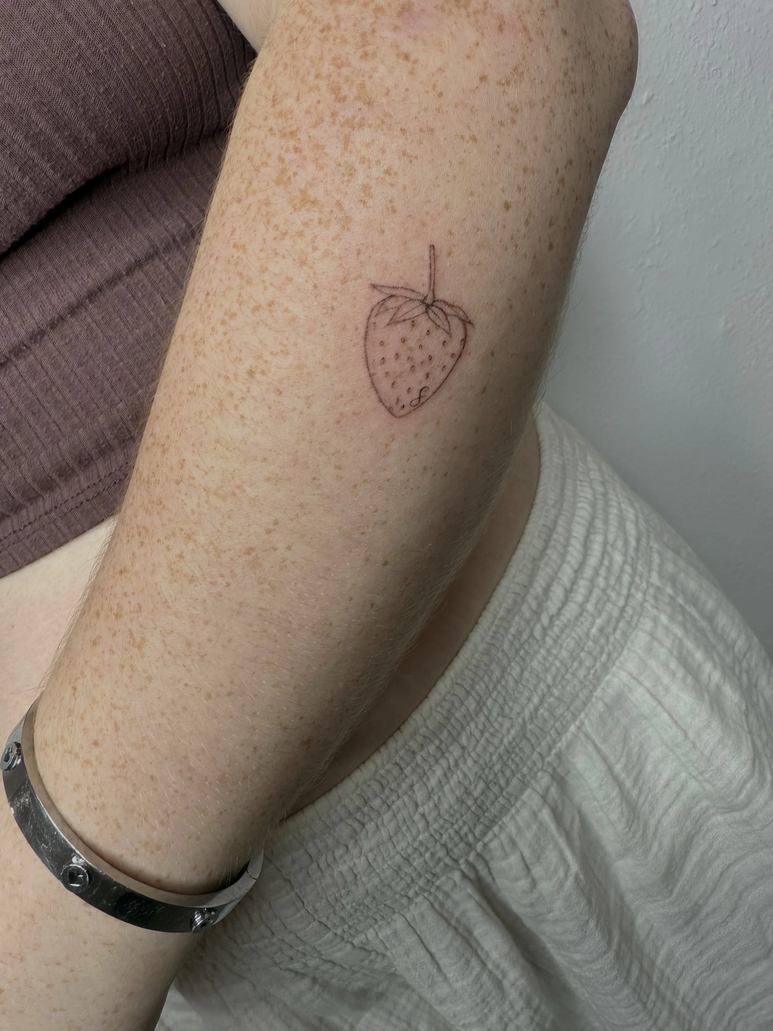 A leg with freckles and a tattoo of a strawberry outline with a small letter 'S' inside, resting on a white textured surface.