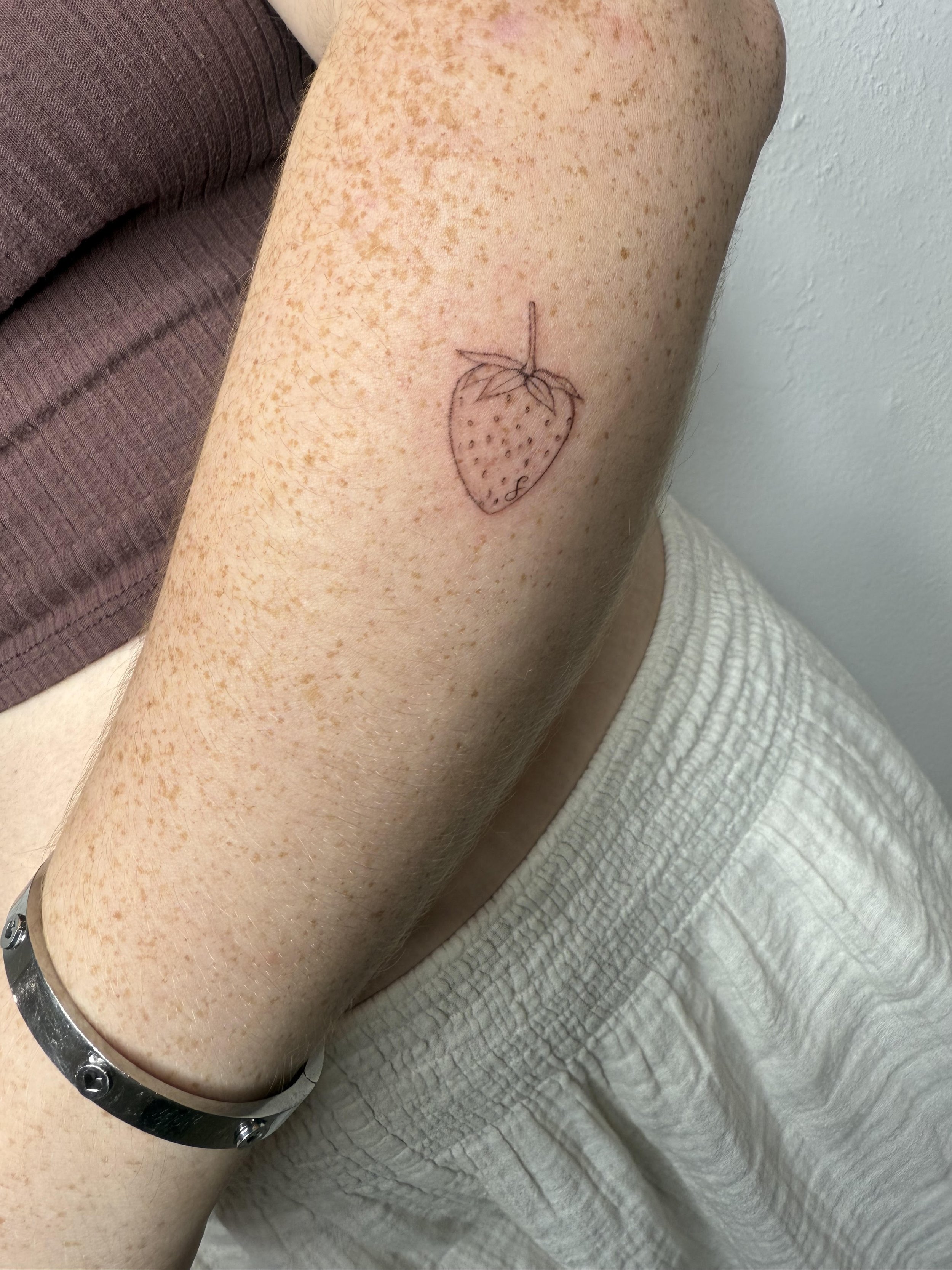 A person with light skin and freckles on their arm has a small sketch of a strawberry tattooed on their upper arm. The strawberry tattoo is a simple outline with small dots representing the seeds and a little stem with leaves at the top.